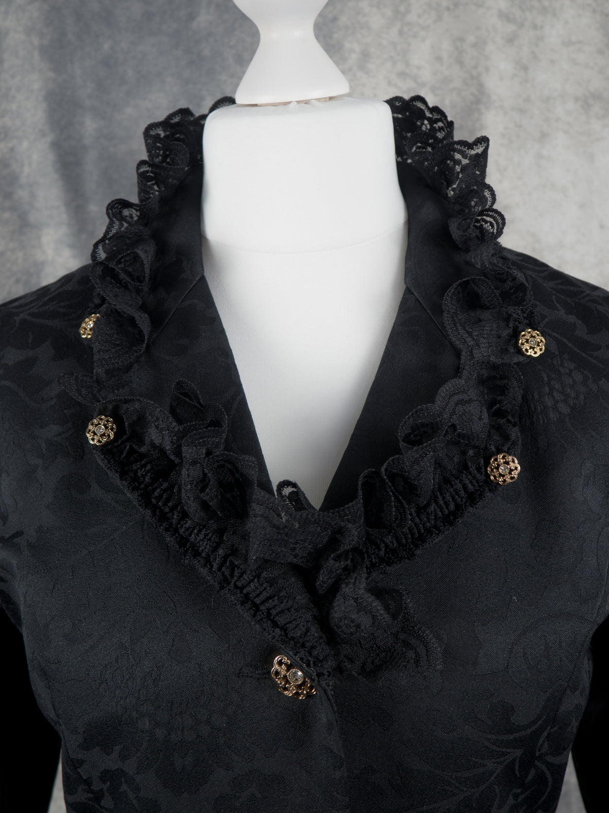 A close-up of a black brocade jacket displayed on a white mannequin torso. The garment features prominent black lace ruffles around the collar and neckline, accented by small gold-tone decorative buttons.