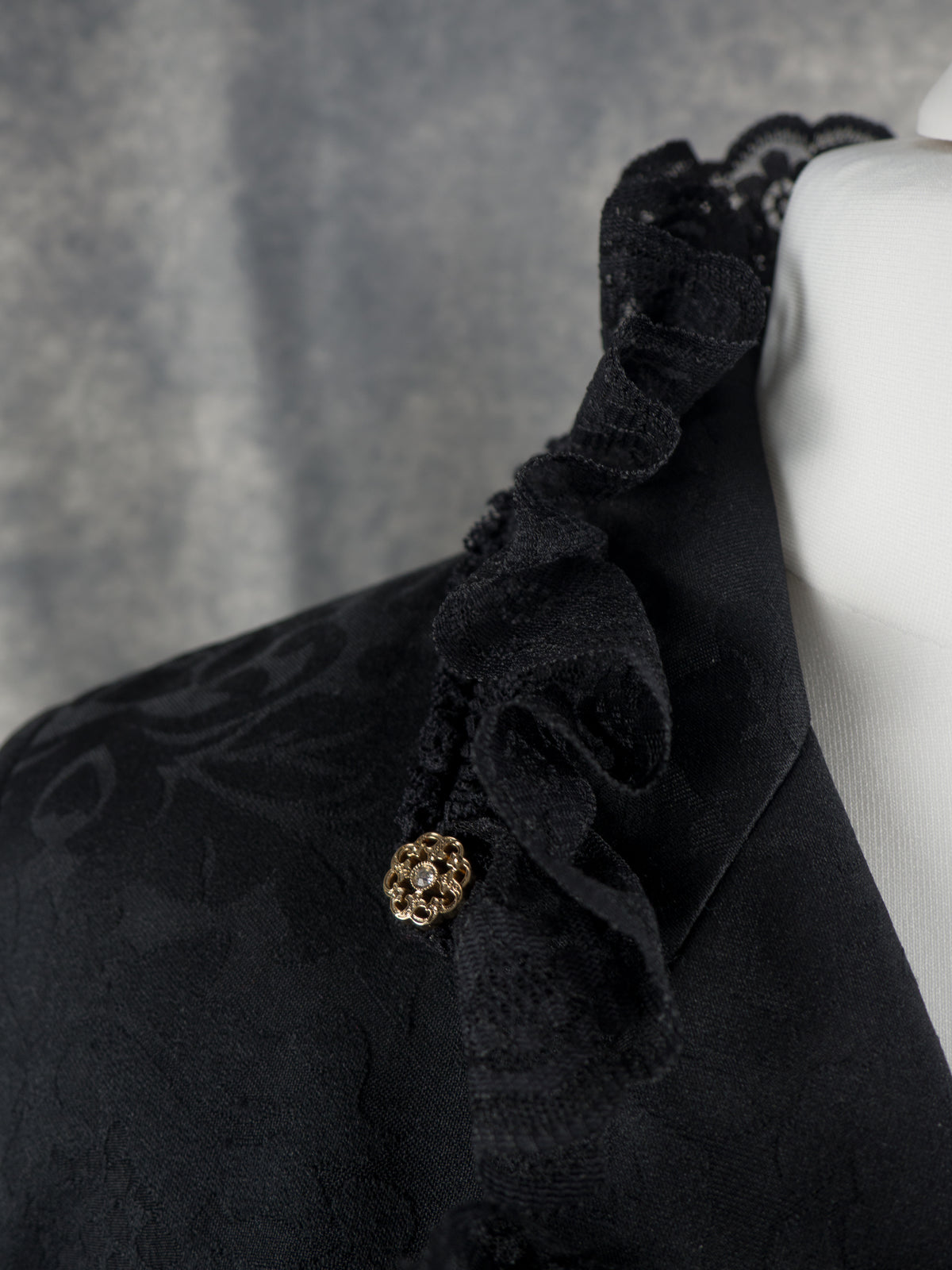 Close-up of a black ruffled lace-trimmed garment on a white mannequin torso, featuring a small gold decorative embellishment and a textured dark gray background.