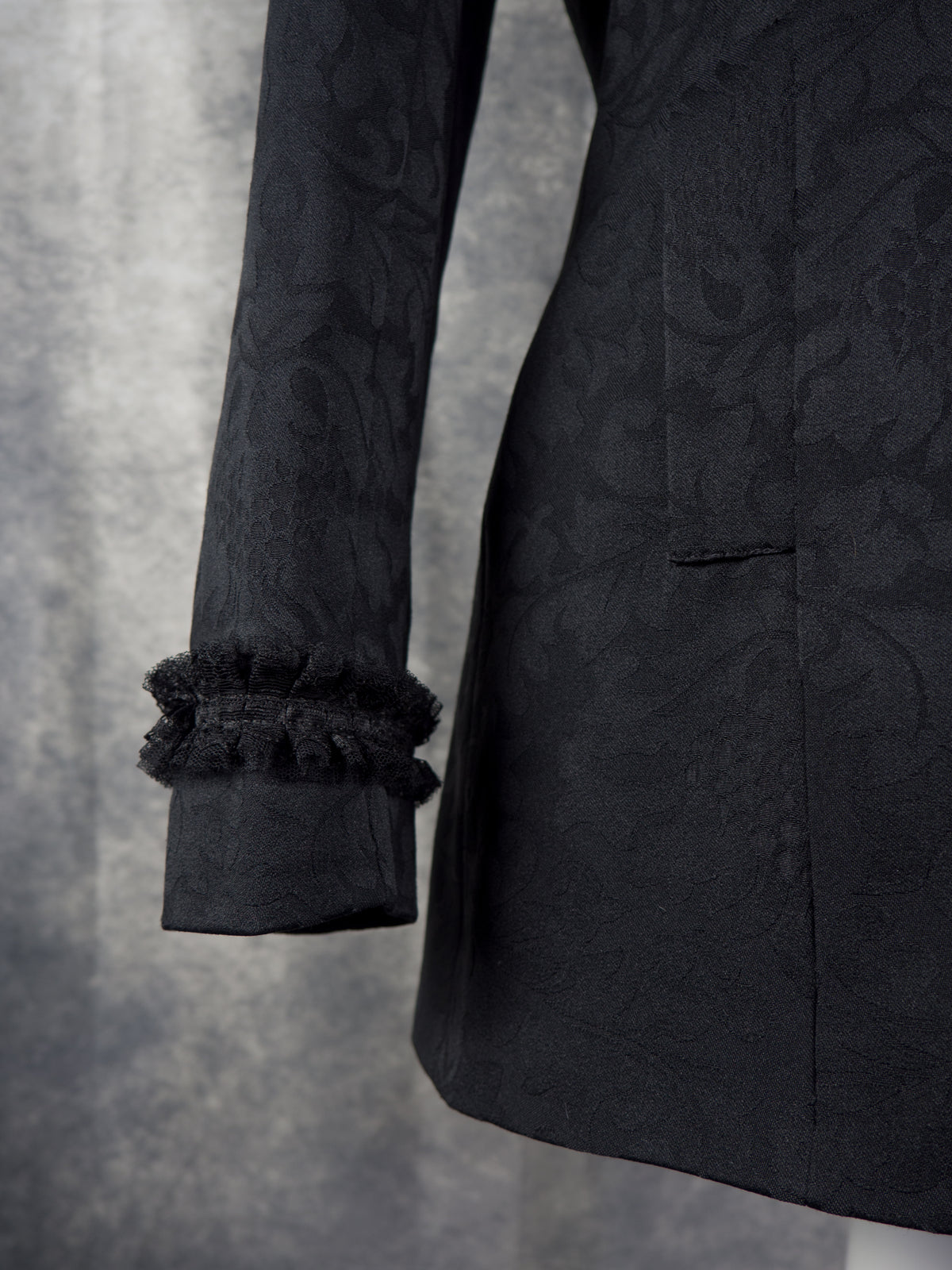 Close-up of a black jacquard-patterned blazer sleeve with a ruffled cuff, set against a textured gray background.