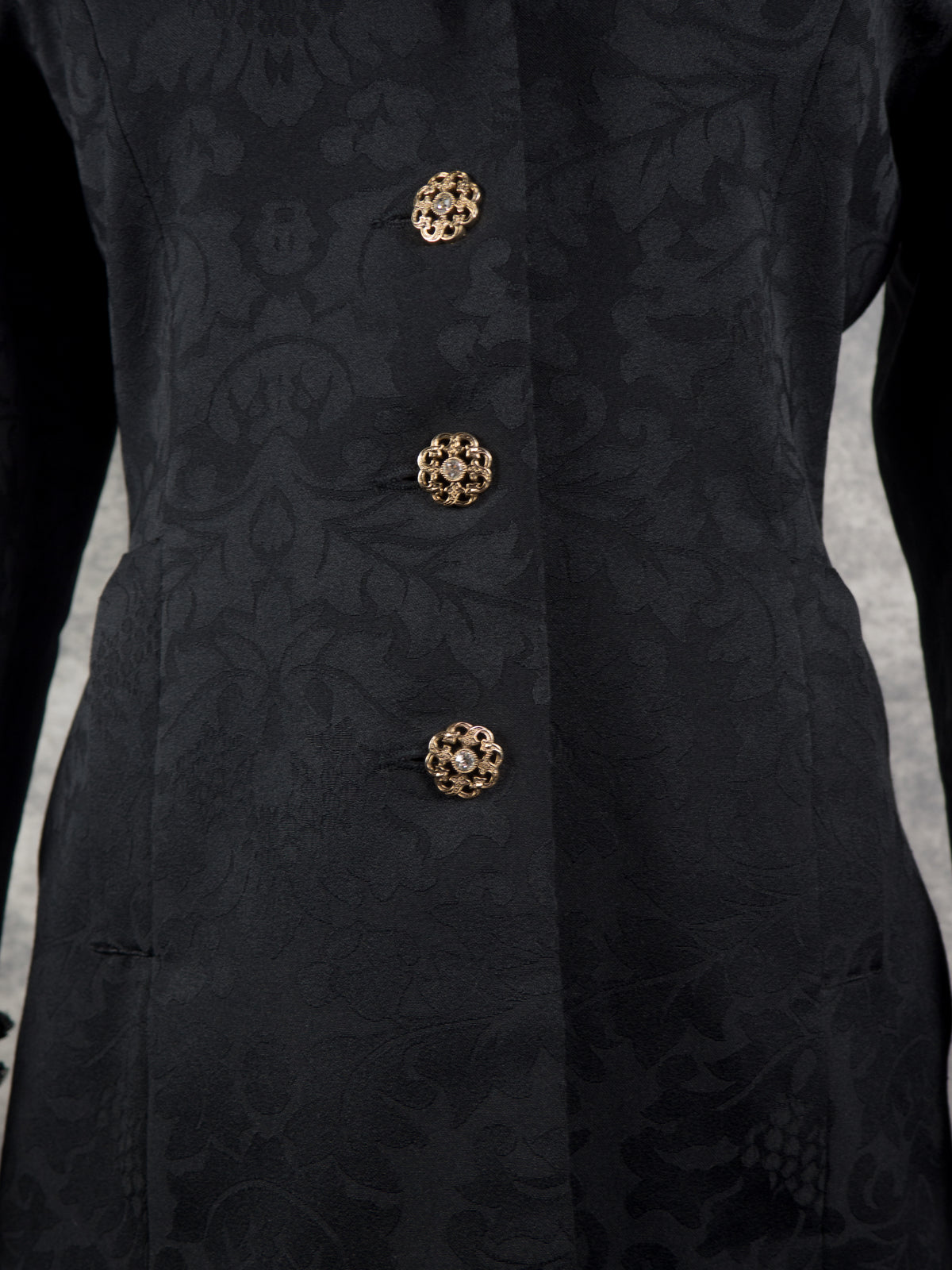 Close-up of a black jacquard jacket with a subtle damask pattern and three ornate gold buttons down the front.