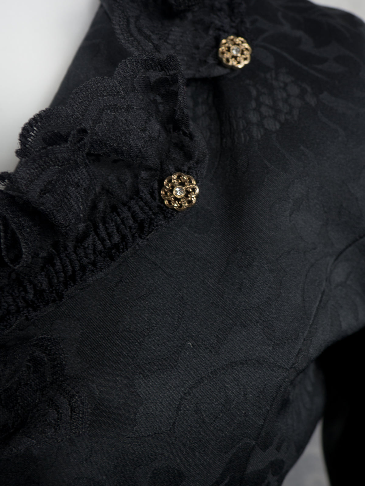 Close-up of a black lace and jacquard fabric garment with ruffled trim and ornate gold-tone decorative buttons.