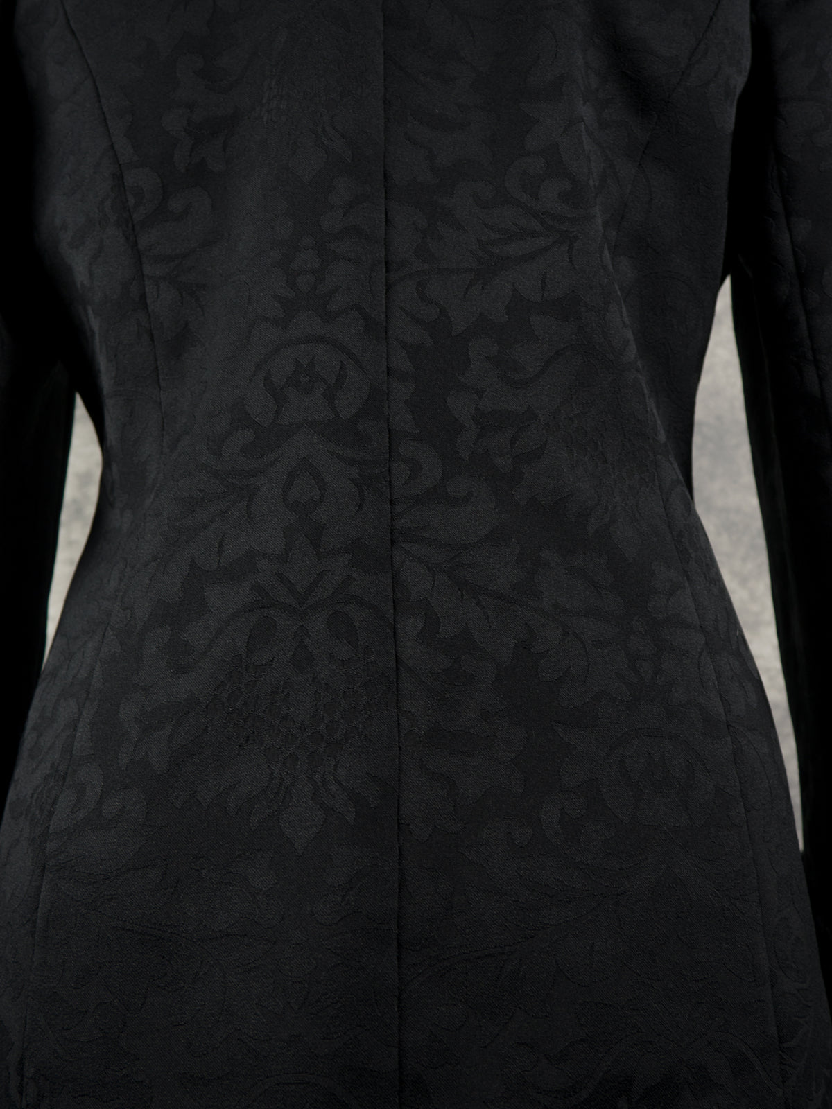 Close-up of a black long-sleeve blazer with a subtle tonal damask pattern and a center back seam.