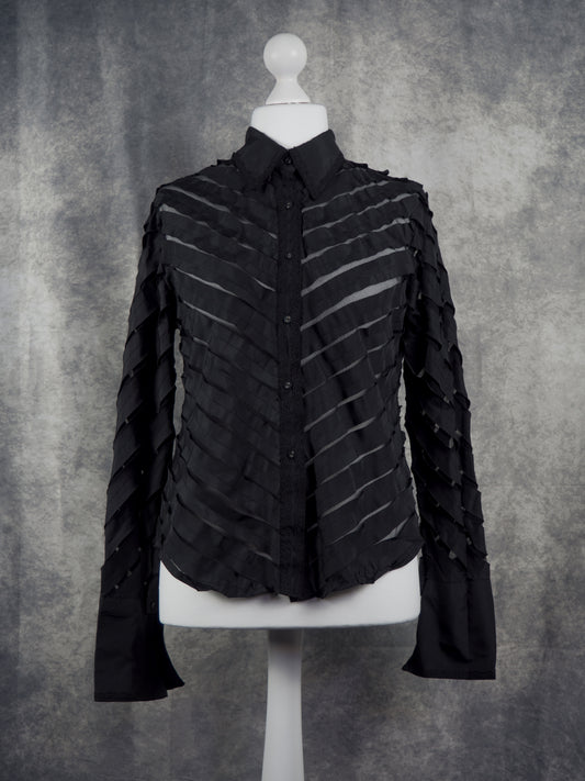 A black long-sleeve button-up shirt with diagonal pleated ruffle detailing, displayed on a white mannequin torso against a textured gray background.