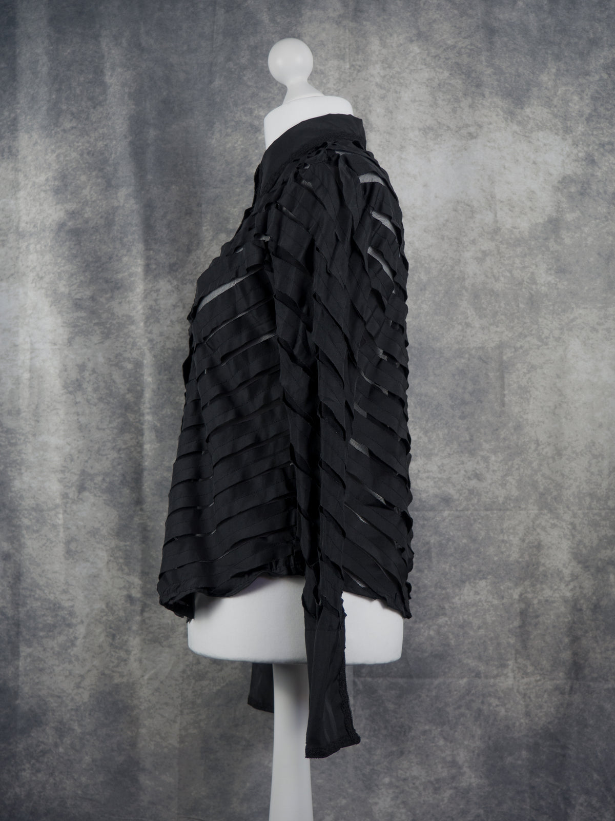 A black long-sleeve blouse with a unique layered, ruffled, and slit design, displayed on a white mannequin torso against a textured gray background.