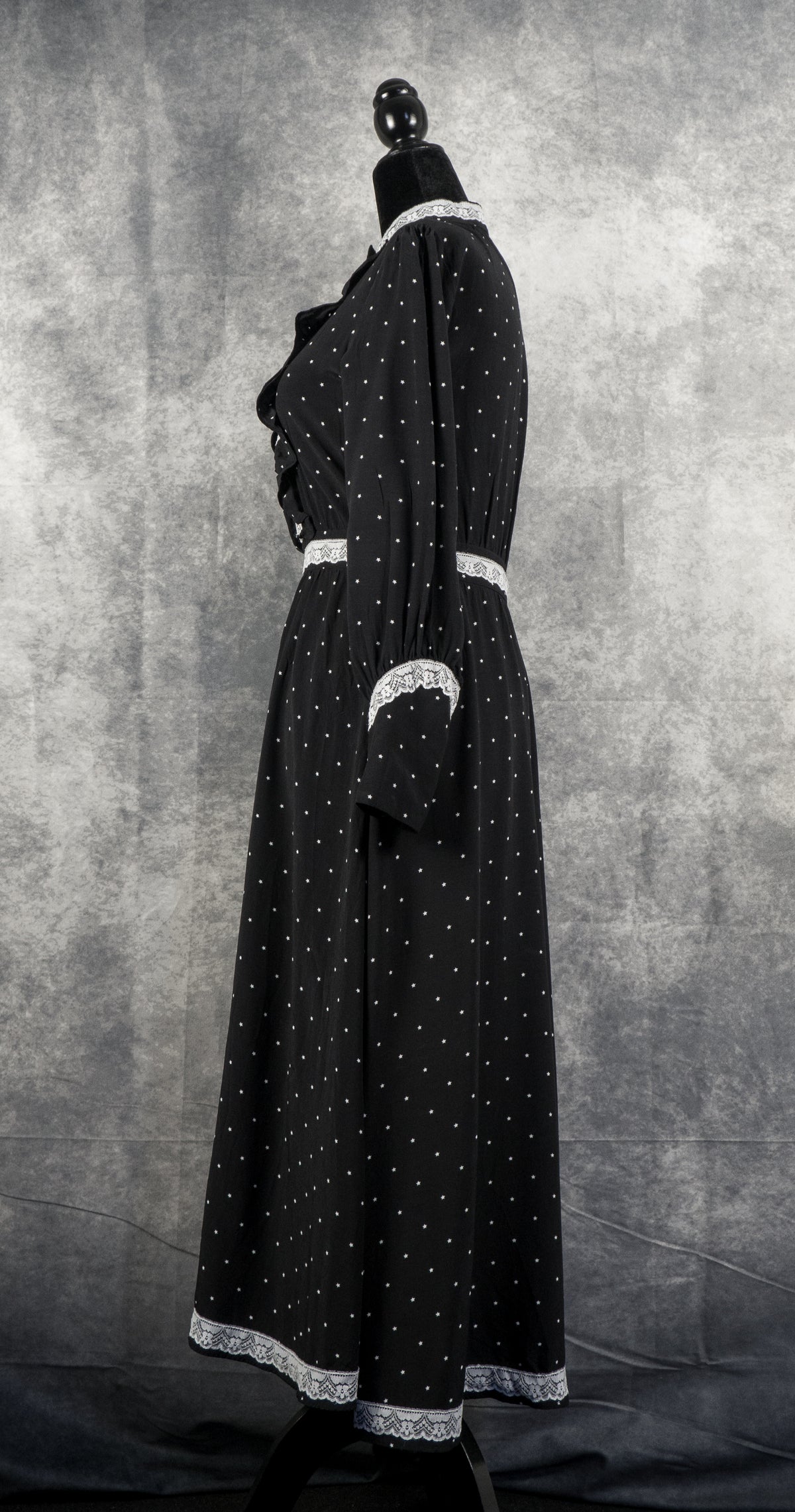 A full-length side view of a vintage-style black maxi dress with long sleeves, displayed on a black mannequin against a textured grey background. The dress features a delicate pattern of small white stars. It is detailed with white lace trim along the high collar, the cuffs, the waistband, and the bottom hem. The dress has long sleeves with ruffled shoulders and buttoned cuffs, creating a classic, prairie-inspired silhouette.