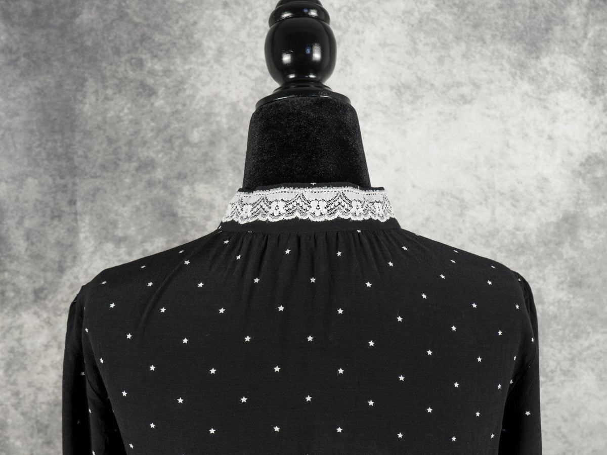 A close-up back view of the upper portion of the black vintage-style dress, displayed on a mannequin against a textured grey background. The image highlights the fine pattern of small white stars scattered across the black fabric, along with gathers at the neckline. A delicate band of white lace trim circles the high collar.