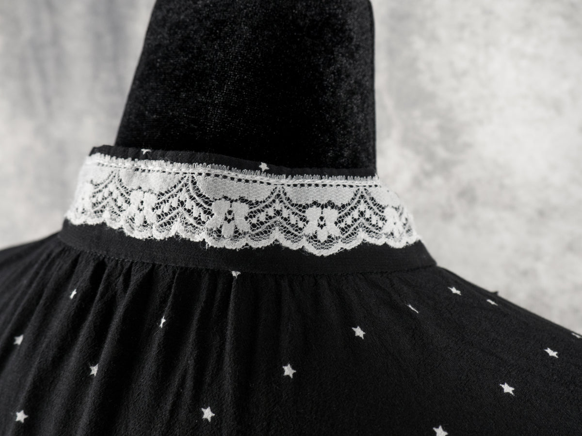 An extreme close-up shot focusing on the back neckline of the black dress with a small white star pattern. The image showcases the intricate details of the white lace trim stitched onto the high collar, as well as the fine gathers in the fabric just below the collar. The dress is displayed on a black mannequin against a soft, textured grey background.