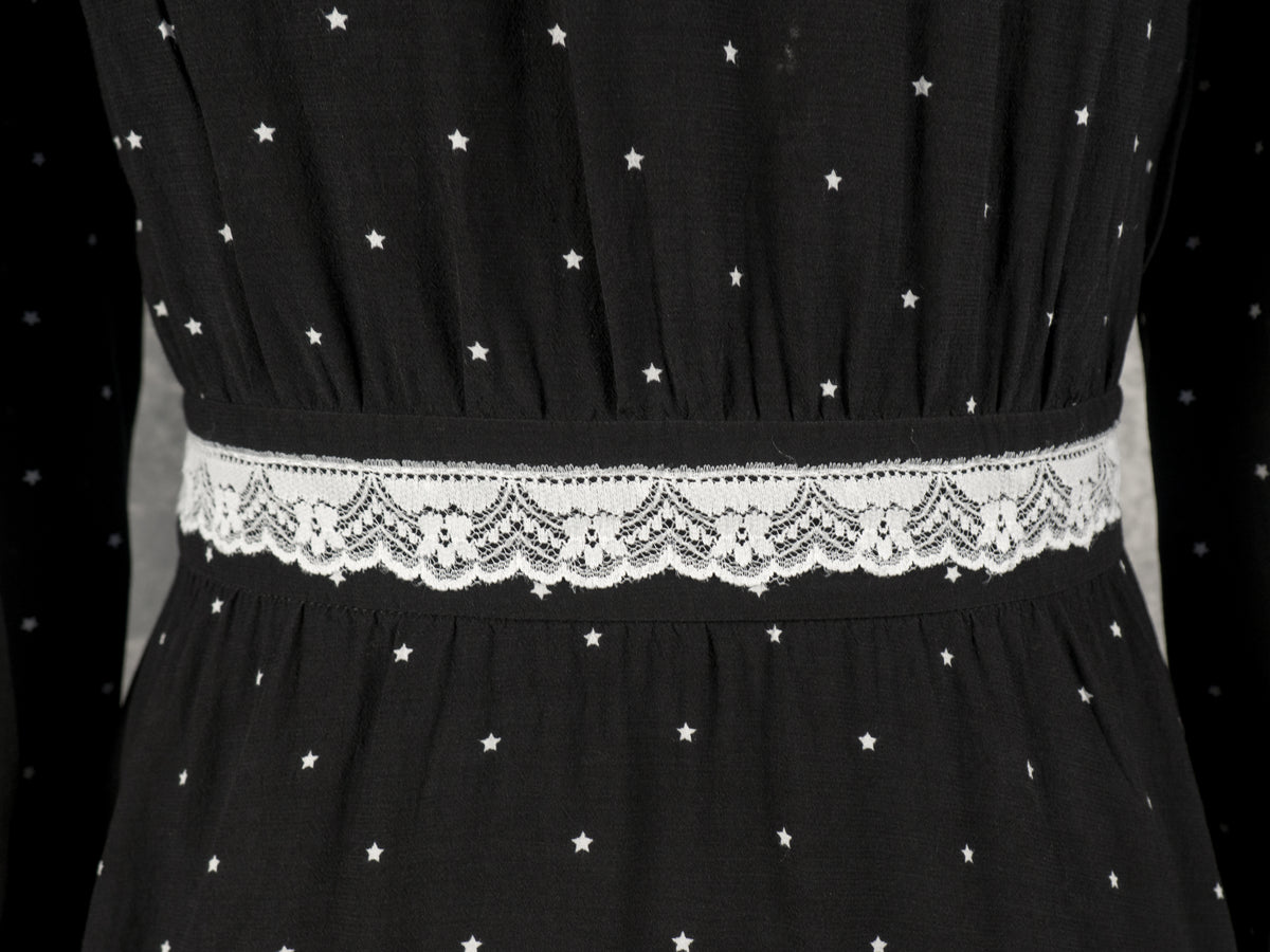 A close-up shot focusing on the waistline of the black dress, highlighting the white lace trim sewn horizontally across the elasticated or gathered waistband. The fabric above and below the waist features the continuous small white star print with subtle vertical gathering details. The long sleeves of the dress are partially visible on both sides against the neutral grey background.