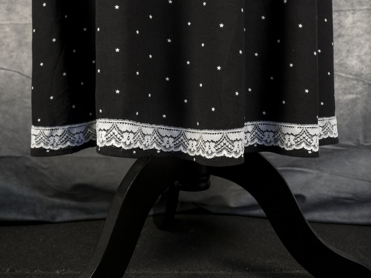 A close-up shot focusing on the bottom hem of the black dress, showing the continuous band of detailed white lace trim running along the edge. The fabric features the pattern of small white stars, and the bottom of the black mannequin stand is visible below the floating hem against a dark floor and textured grey backdrop.