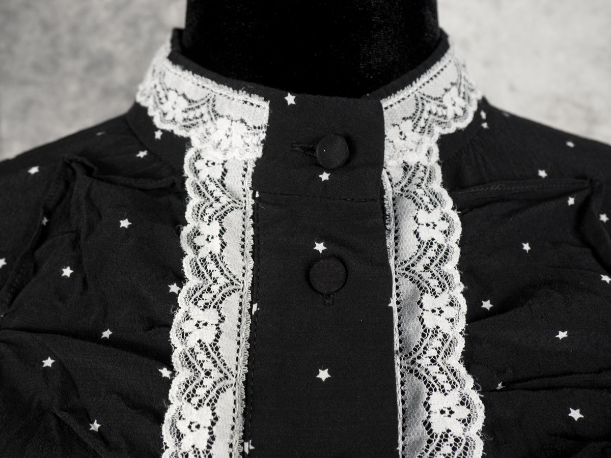 A detailed close-up front view of the dress's collar and placket. The image shows the high band collar adorned with white lace trim, which extends down vertically on both sides of the central fabric placket. Two black fabric-covered buttons are fastened along the placket, set against the black fabric with the small white star pattern and subtle gathering at the chest.