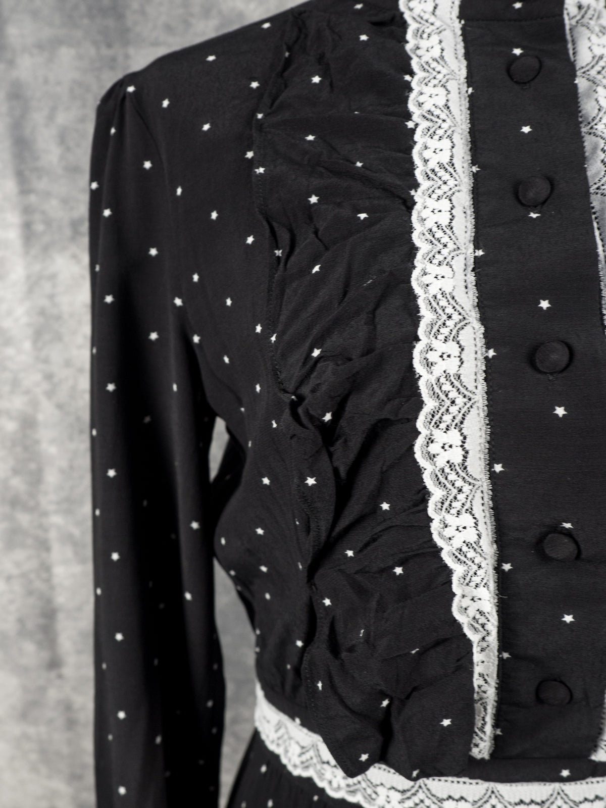 A close-up shot focusing on the front bodice and ruffle detail of the black dress. The image highlights a vertical ruffle panel running alongside the vertical white lace trim and button placket, which features several black fabric-covered buttons. The fine pattern of small white stars covers the slightly wrinkled or textured fabric, and a portion of the long sleeve and lace-trimmed waistband are also visible.