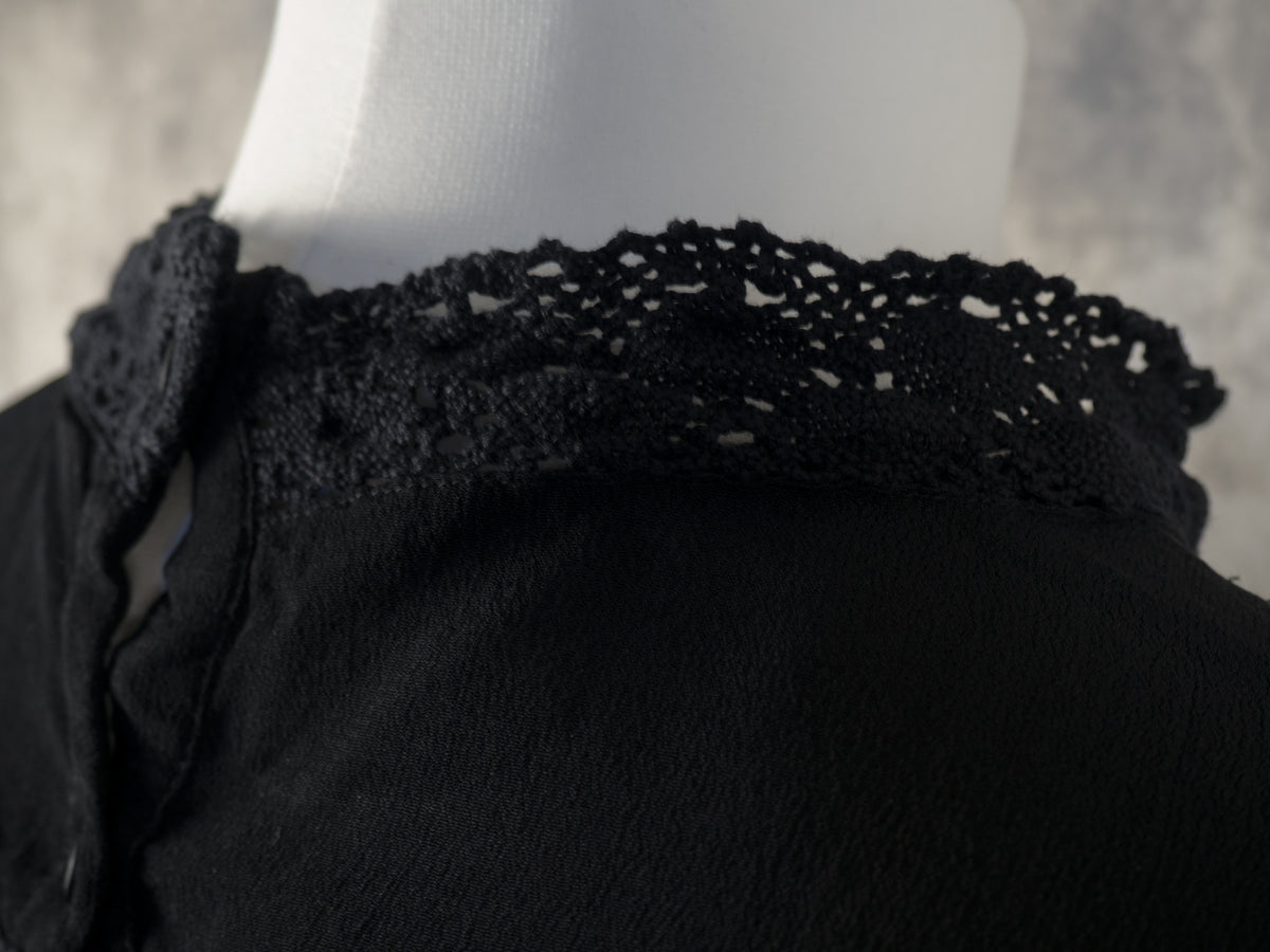 A close-up view of the high neckline on a black dress, highlighting the delicate crocheted lace collar that wraps around the neck. The image captures the intricate openwork pattern of the lace trim and the soft texture of the black fabric on a white mannequin, against a grey background.