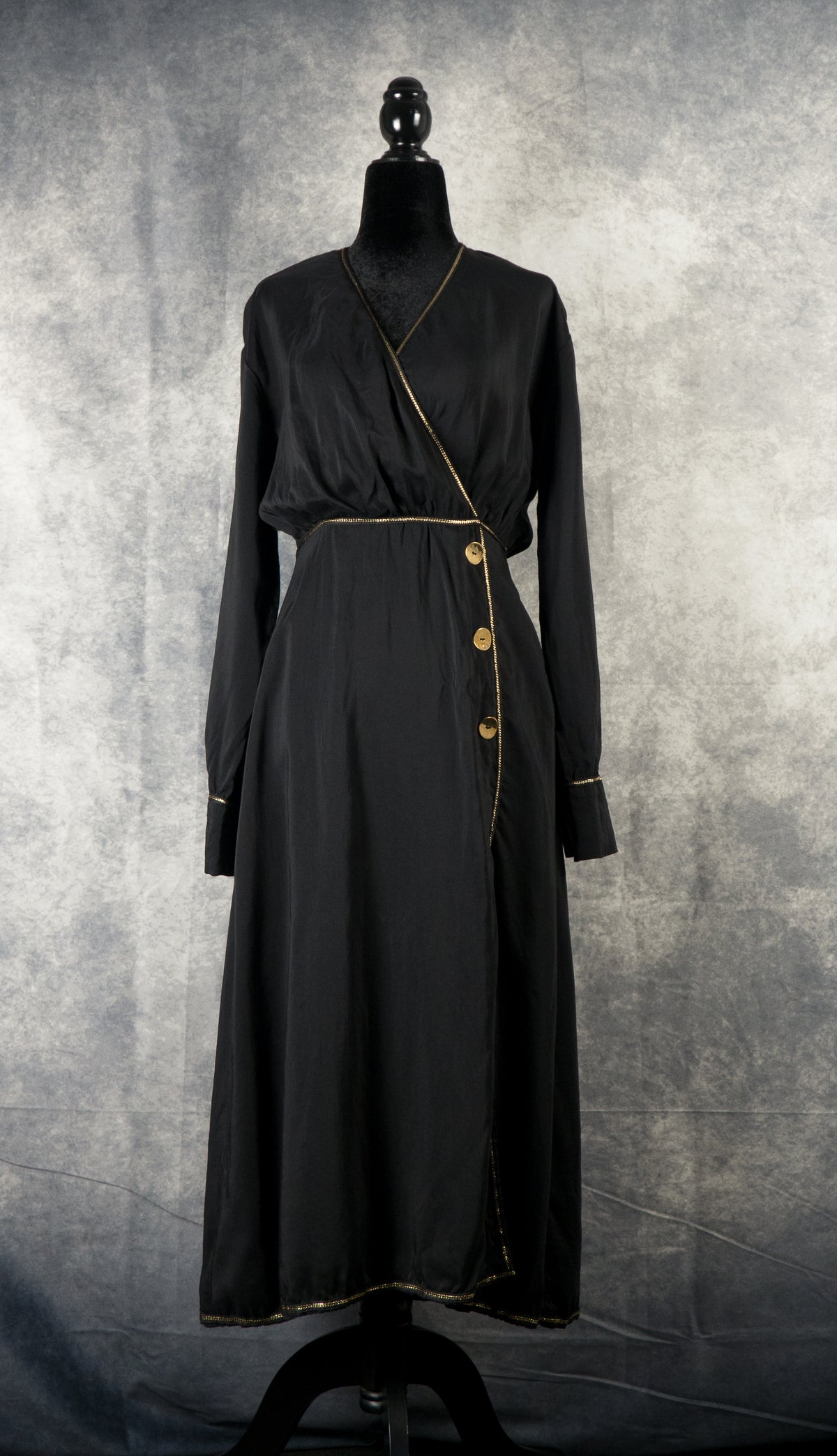 A long-sleeved, black maxi wrap dress displayed on a black mannequin against a textured gray background. The dress features a V-neckline, a wrap-around bodice secured with three gold decorative buttons on the left hip, and an asymmetric front slit. Thin gold trim accentuates the neckline, waist, cuffs, and hemline.