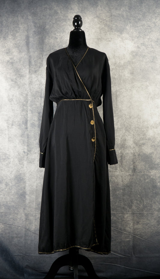 A long-sleeved, black maxi wrap dress displayed on a black mannequin against a textured gray background. The dress features a V-neckline, a wrap-around bodice secured with three gold decorative buttons on the left hip, and an asymmetric front slit. Thin gold trim accentuates the neckline, waist, cuffs, and hemline.