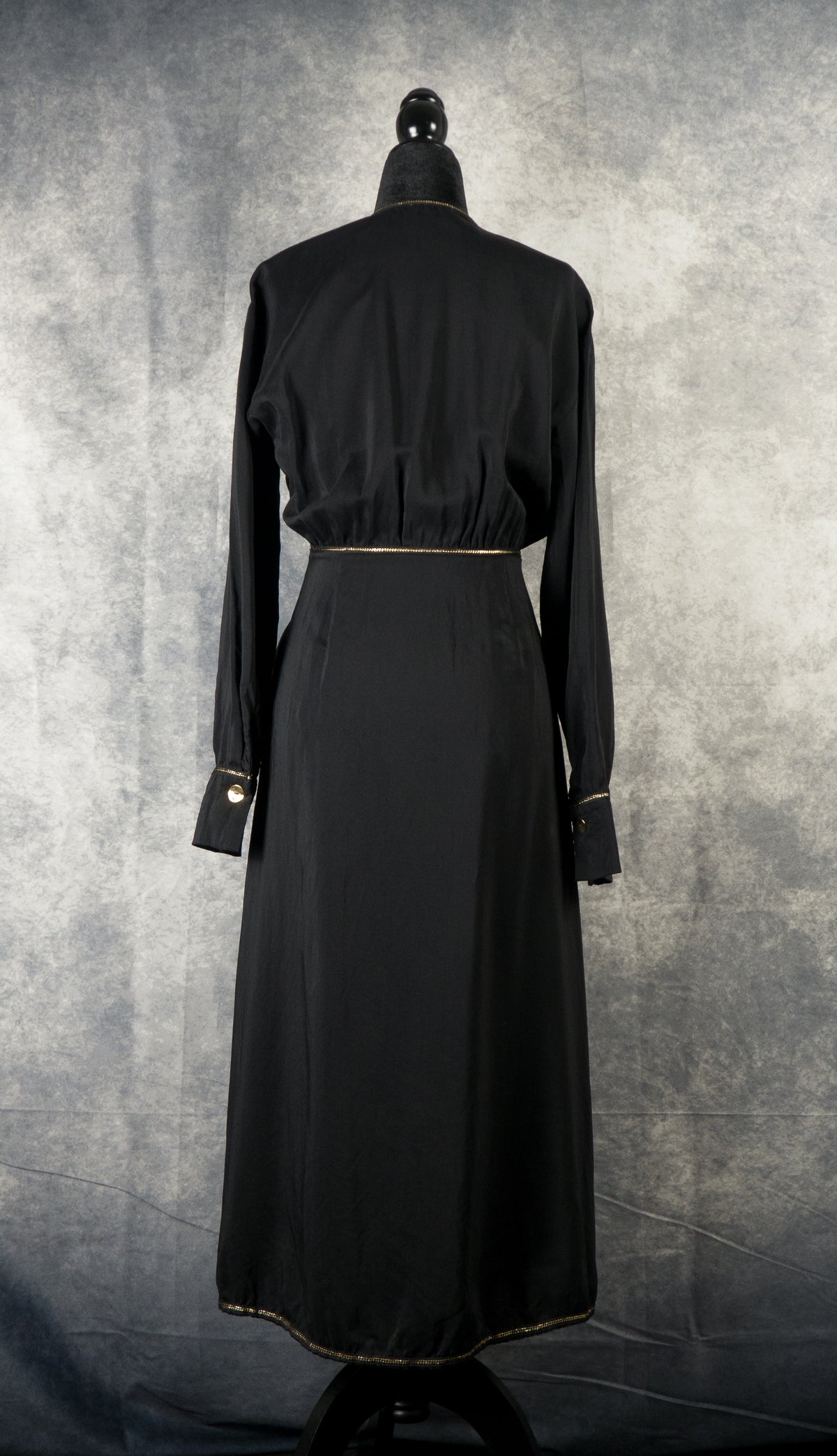 The back view of a long-sleeved, black maxi dress displayed on a black mannequin against a textured gray background. The dress features a gathered elastic waist detailed with a thin gold trim that continues from the front. The long sleeves end in structured cuffs, each accented with a small gold button and gold trim. A subtle gold line also edges the bottom hem of the straight-cut skirt.