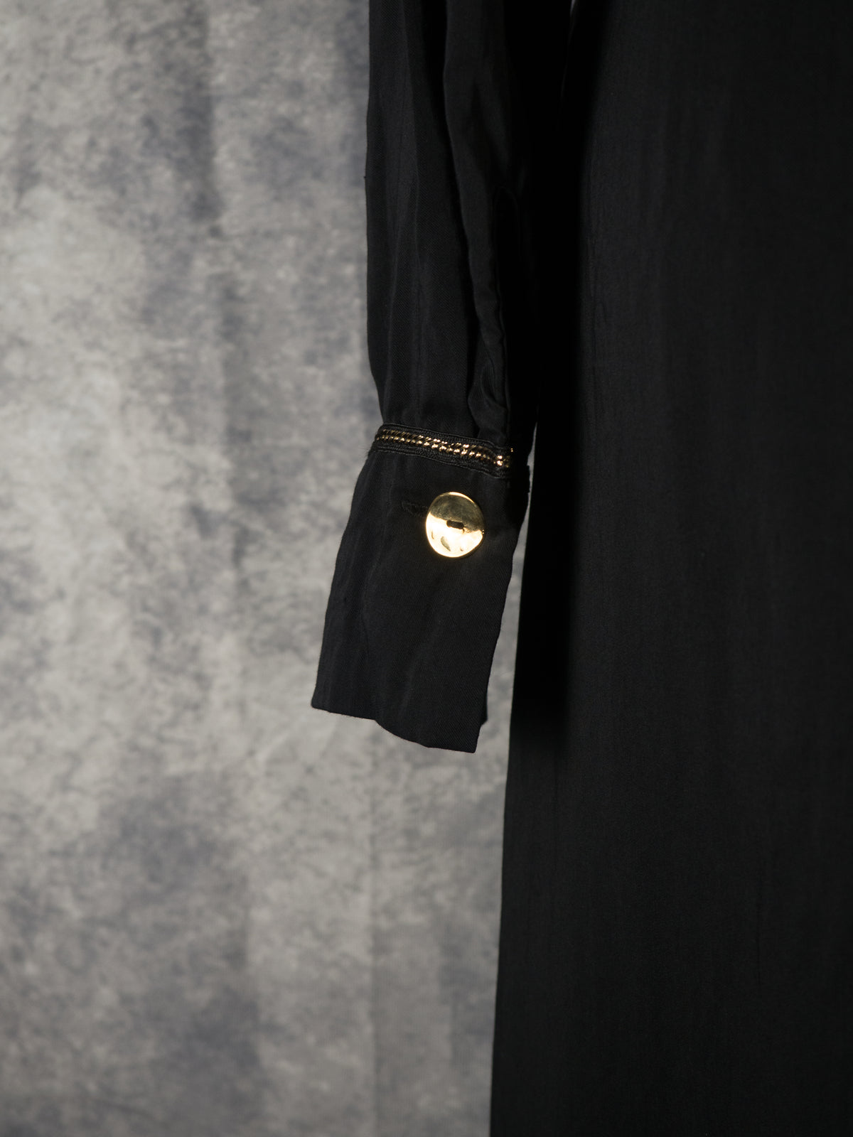 A close-up shot of the long sleeve cuff on a black dress. The cuff is fastened with a prominent, shiny round gold button and is detailed with a delicate metallic gold chain trim running along its upper seam. The background is a textured grey backdrop.
