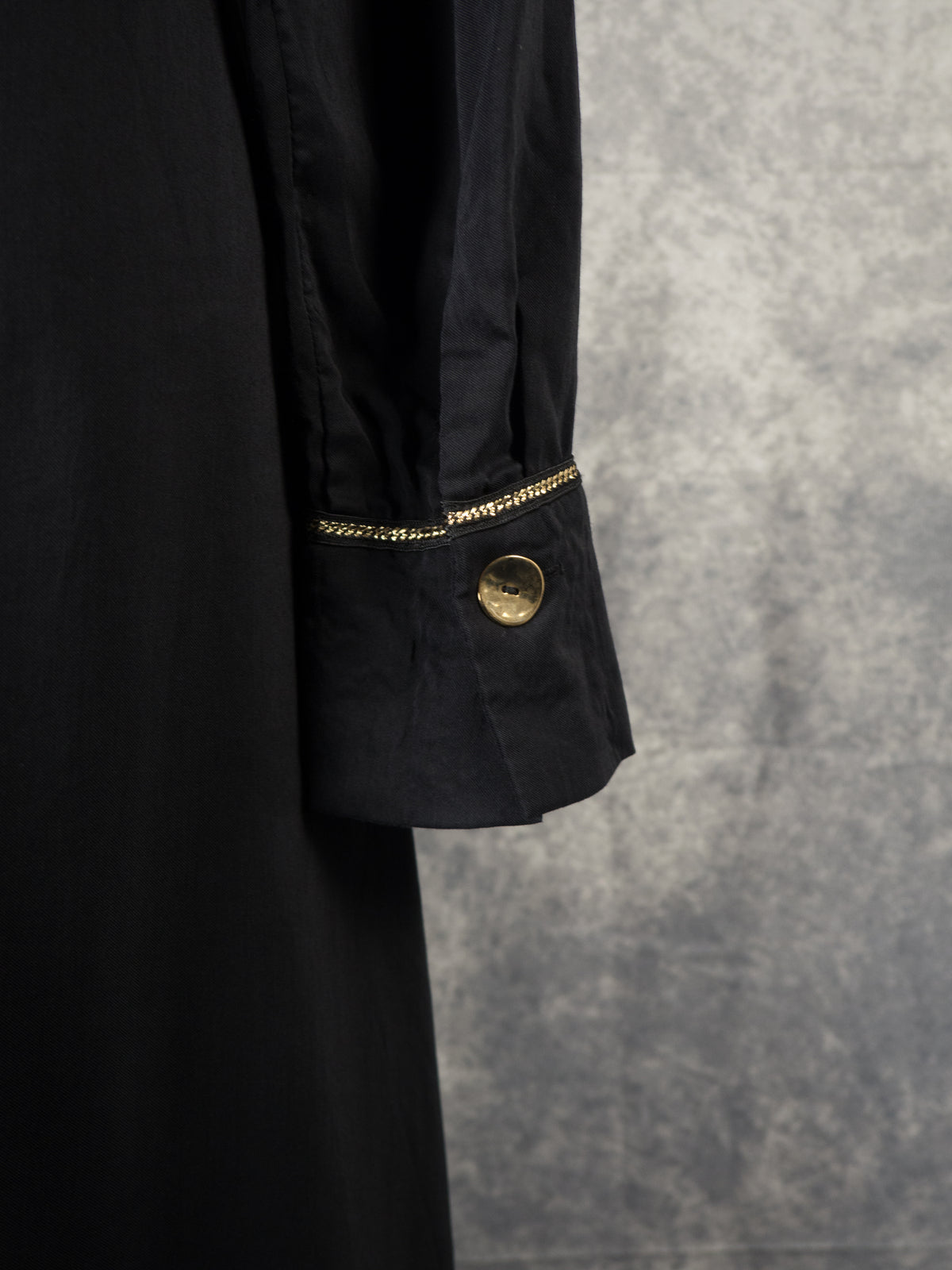 A close-up shot of the long sleeve cuff on a black dress. The cuff is fastened with a prominent, shiny round gold button and is detailed with a delicate metallic gold chain trim running along its upper seam. The background is a textured grey backdrop.