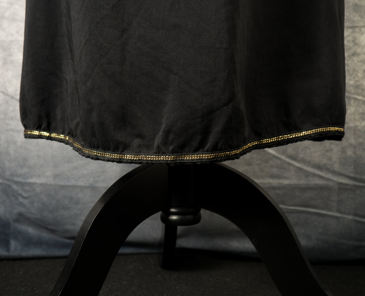 A close-up shot of the lower hem of a black maxi dress. A delicate, metallic gold chain trim runs along the entire edge of the slightly gathered hemline. The black wooden base of the mannequin stand is visible directly underneath against a textured gray background.