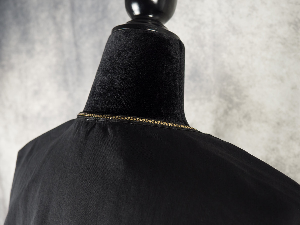 A close-up shot of the back neckline of a black dress on a mannequin. A delicate, metallic gold chain trim runs continuously along the stitched collar edge. The background features a soft, textured grey surface.