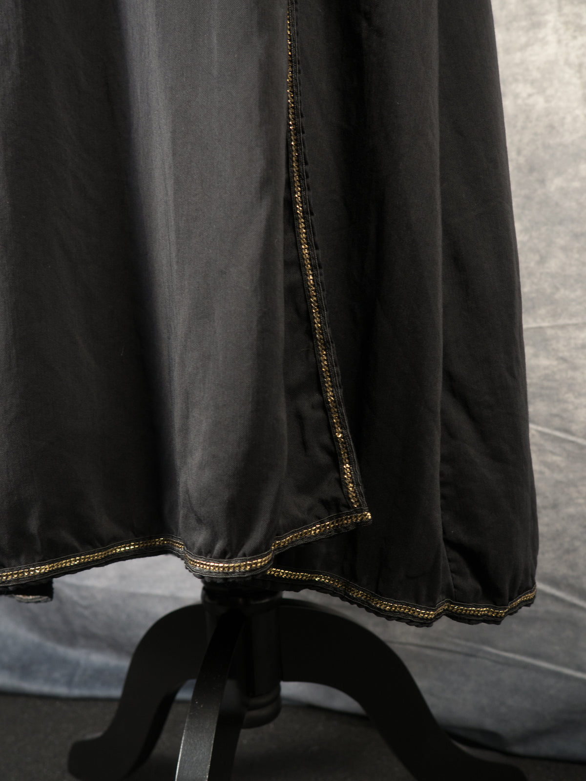 A close-up shot of the lower section of a black wrap dress, showcasing an overlapping wrap detail that creates an asymmetric front slit. A thin, metallic gold chain trim runs along the vertical edge of the opening and extends continuously across the entire bottom hemline. The black wooden base of the mannequin stand is partially visible at the bottom.