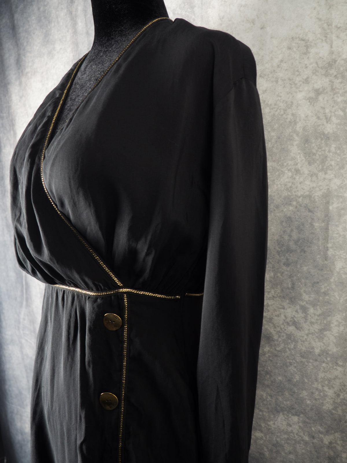 A side-profile close-up shot of a black wrap dress on a mannequin. It emphasizes the draped, crossover wrap bodice with a deep V-neckline and long sleeve. A delicate metallic gold chain trim outlines the neckline and the gathered waist. Two large, round gold buttons are visible on the front panel against a textured gray background.
