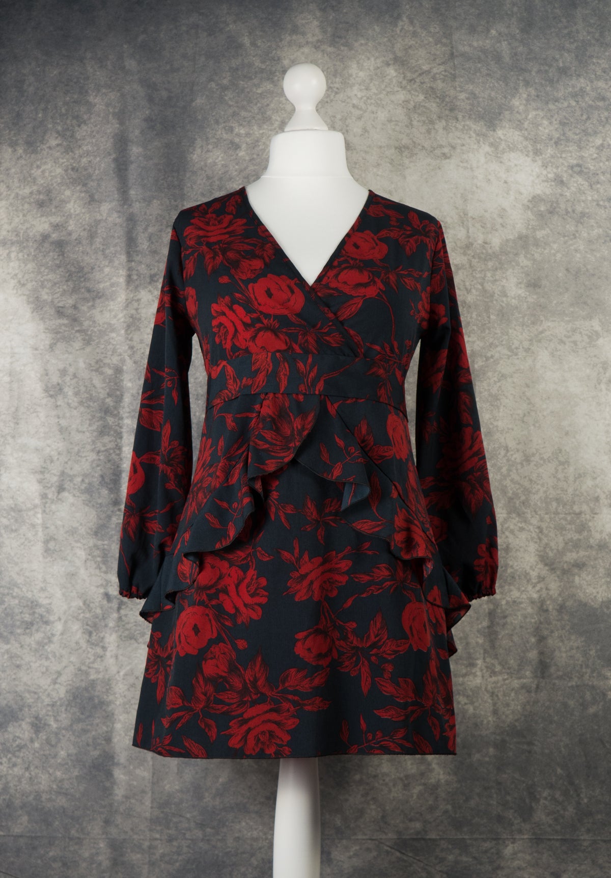 A long-sleeved mini smock dress in black with a vibrant dark red rose floral pattern. It features a deep V-neckline, long sleeves with elasticated cuffs, and a defined waist panel. The skirt and waist are detailed with layered, cascading ruffles that add volume and movement. Displayed on a white mannequin against a textured grey studio background.