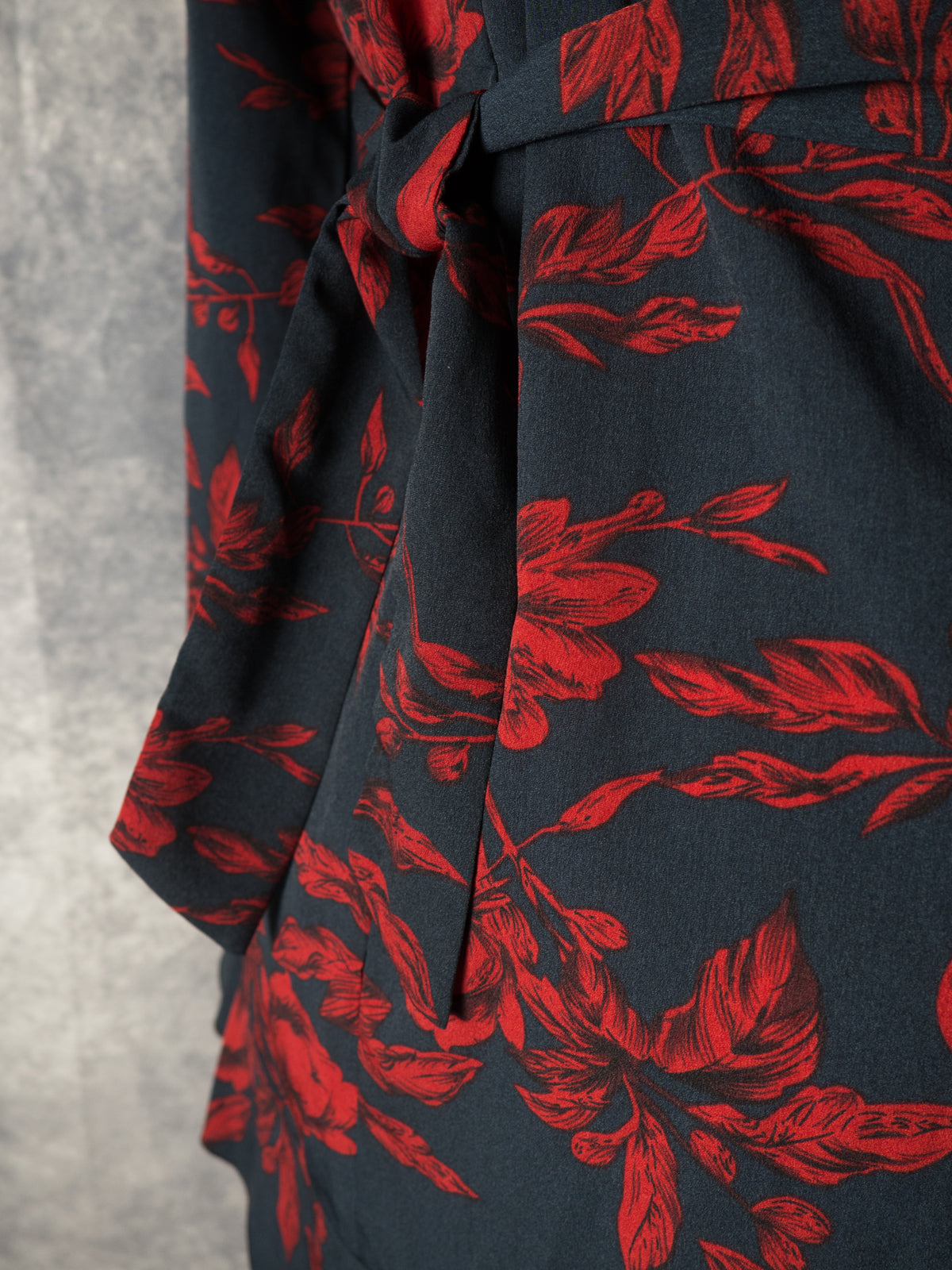 Close-up detail of the waist area of a black dress featuring a dark red floral print. The image highlights a matching fabric tie belt knotted gently at the waist, showing the rich texture of the fabric and leaf pattern against a textured grey background.