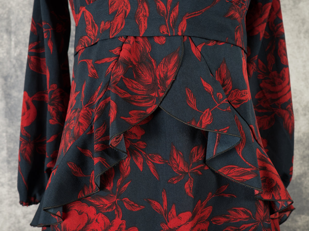 Detailed close-up of the front bodice and skirt of a black dress with a dark red rose print. The image highlights the overlapping, structured ruffles cascading down from the waist panel, creating a layered effect. The intricate floral design and fabric texture are clearly visible against a grey studio backdrop.