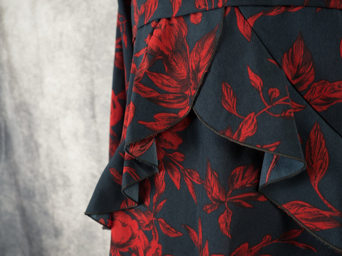 A close-up angled shot focusing on the ruffled detailing of a black dress with a dark red floral pattern. The image showcases the overlapping, layered edges of the cascading ruffles below the waistline, emphasizing the flow and structure of the fabric against a soft grey background.