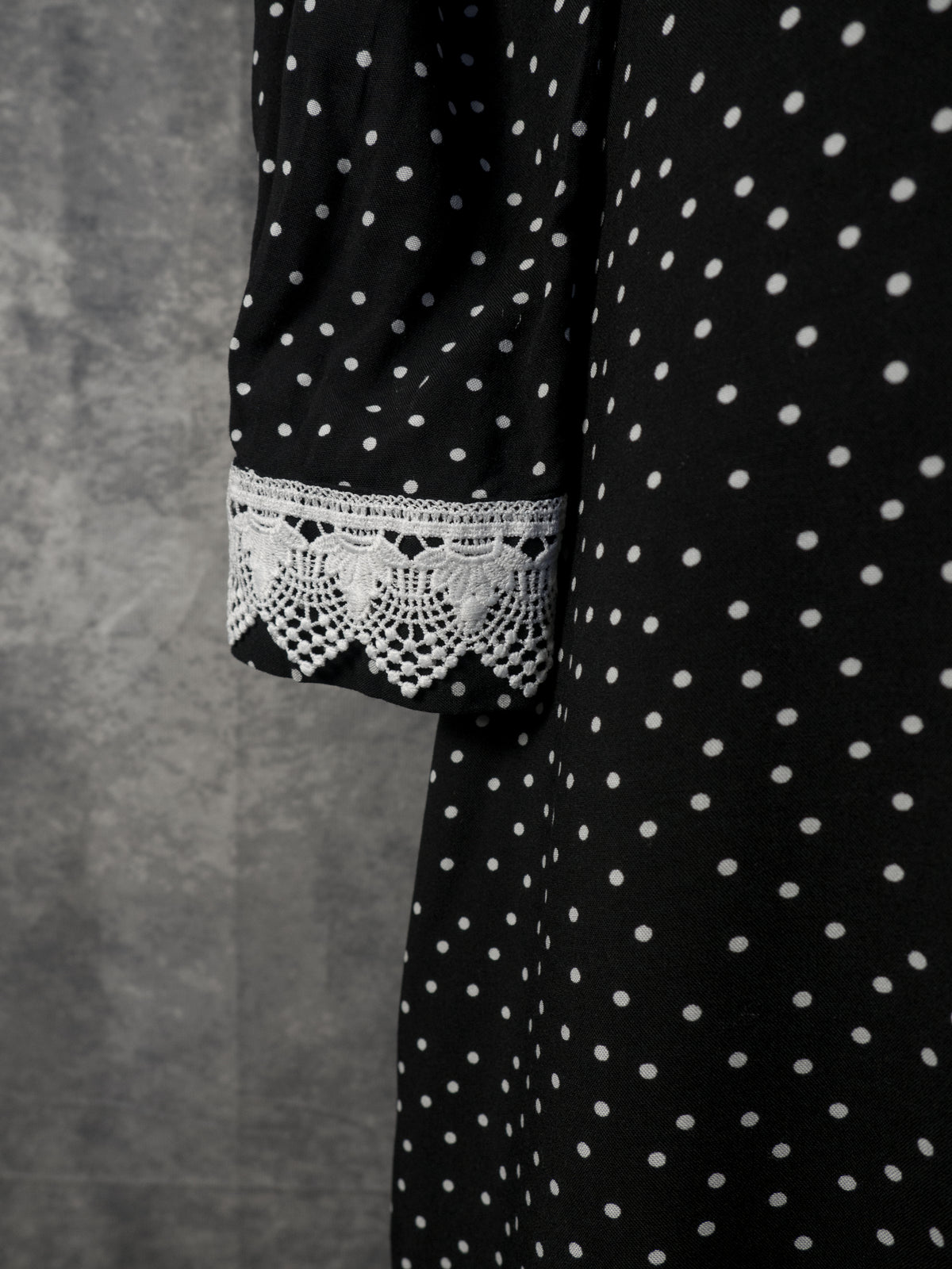 Close-up of a black long-sleeved garment featuring a white polka dot pattern. The sleeve cuff is adorned with a prominent white lace trim showcasing an intricate, scalloped crochet pattern. The background is a textured grey wall.