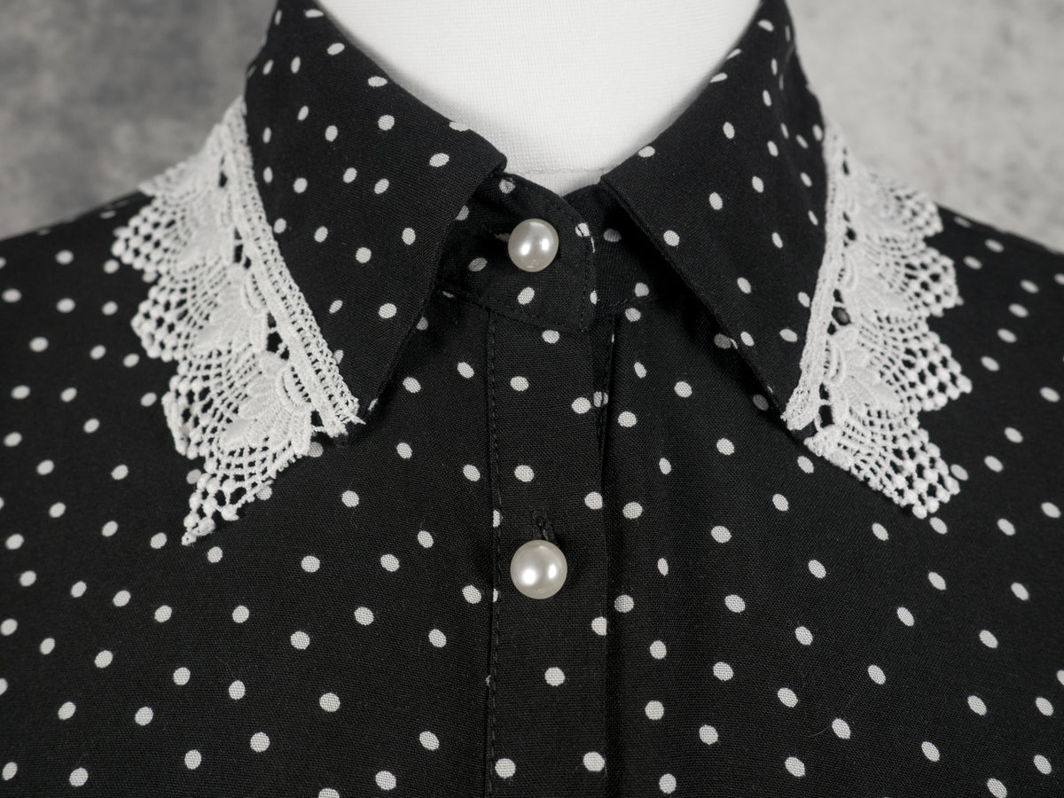 Close-up of the collar and neckline of a black button-up blouse with a white polka dot pattern. The pointed shirt collar features a delicate white crochet lace trim along the edges. The blouse is fastened with round, white pearl-like buttons, and the background is a textured grey wall.
