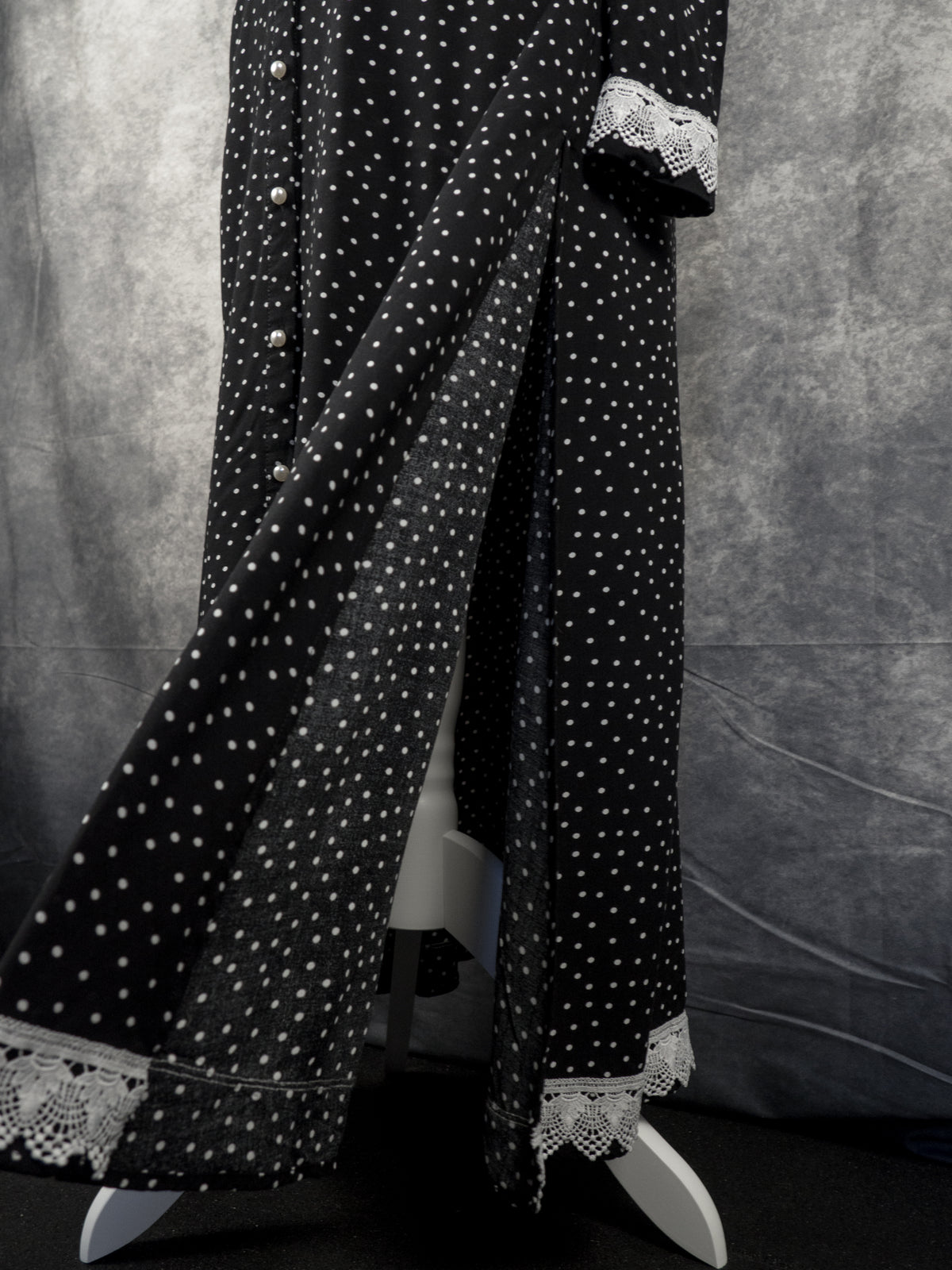 A full-length black dress with a white polka dot pattern displayed on a mannequin. The dress features white pearl-like buttons down the front, a high slit opening, and white crochet lace trim along the cuffs and bottom hem. The background consists of a textured grey fabric backdrop.