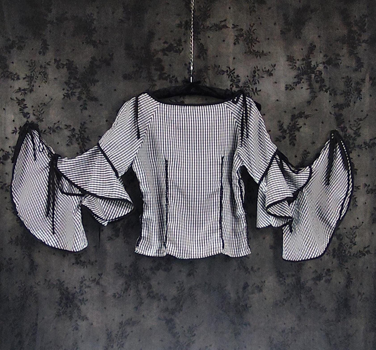 Back view of a black and white vertically striped blouse with dramatic ruffled bell sleeves, hanging on a black hanger against a dark, textured gray background.
