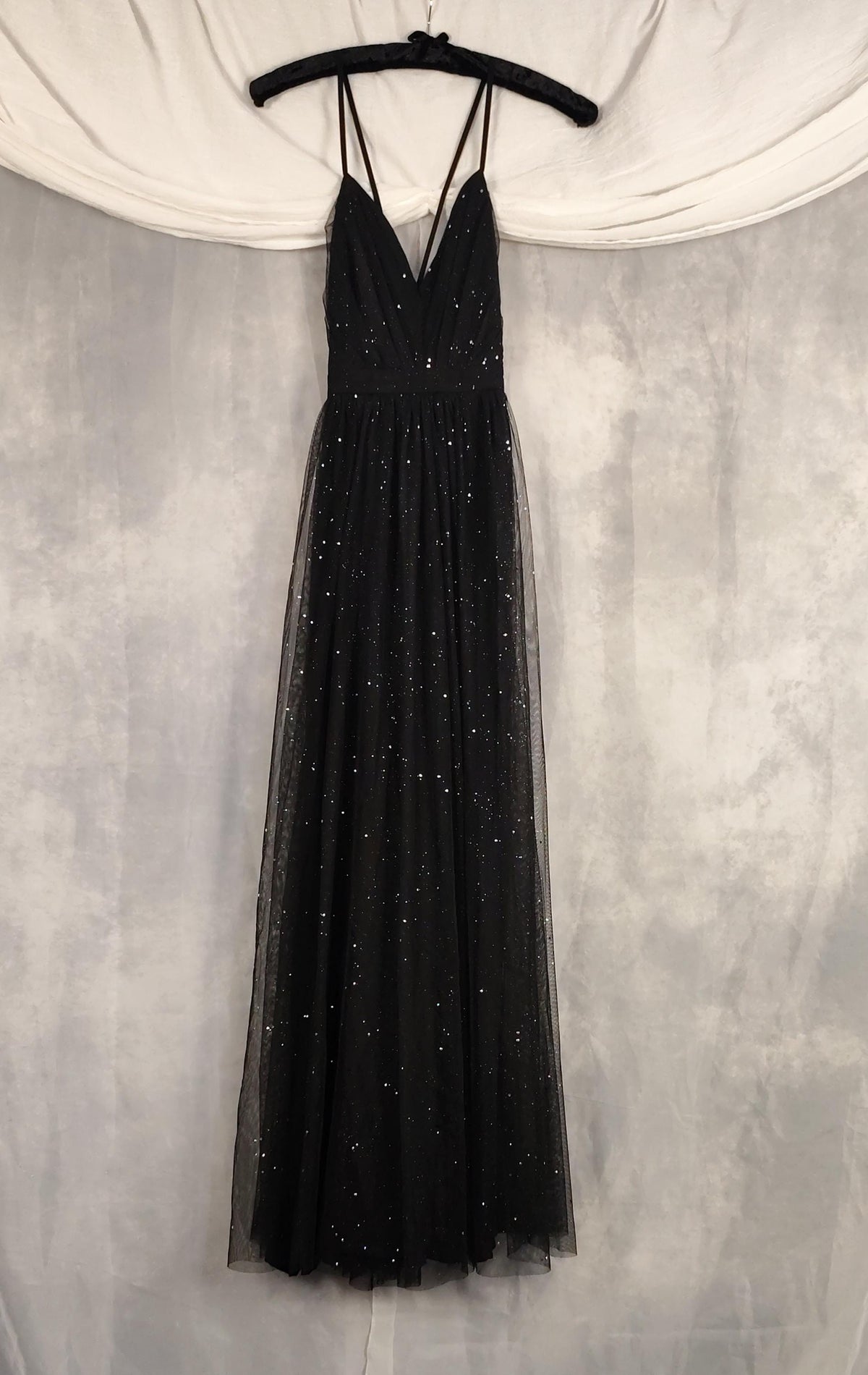 A black sleeveless tulle maxi dress with thin spaghetti straps and a V-neckline, decorated with scattered silver sequins, hanging on a black hanger against a gray textured backdrop with a white draped fabric at the top.