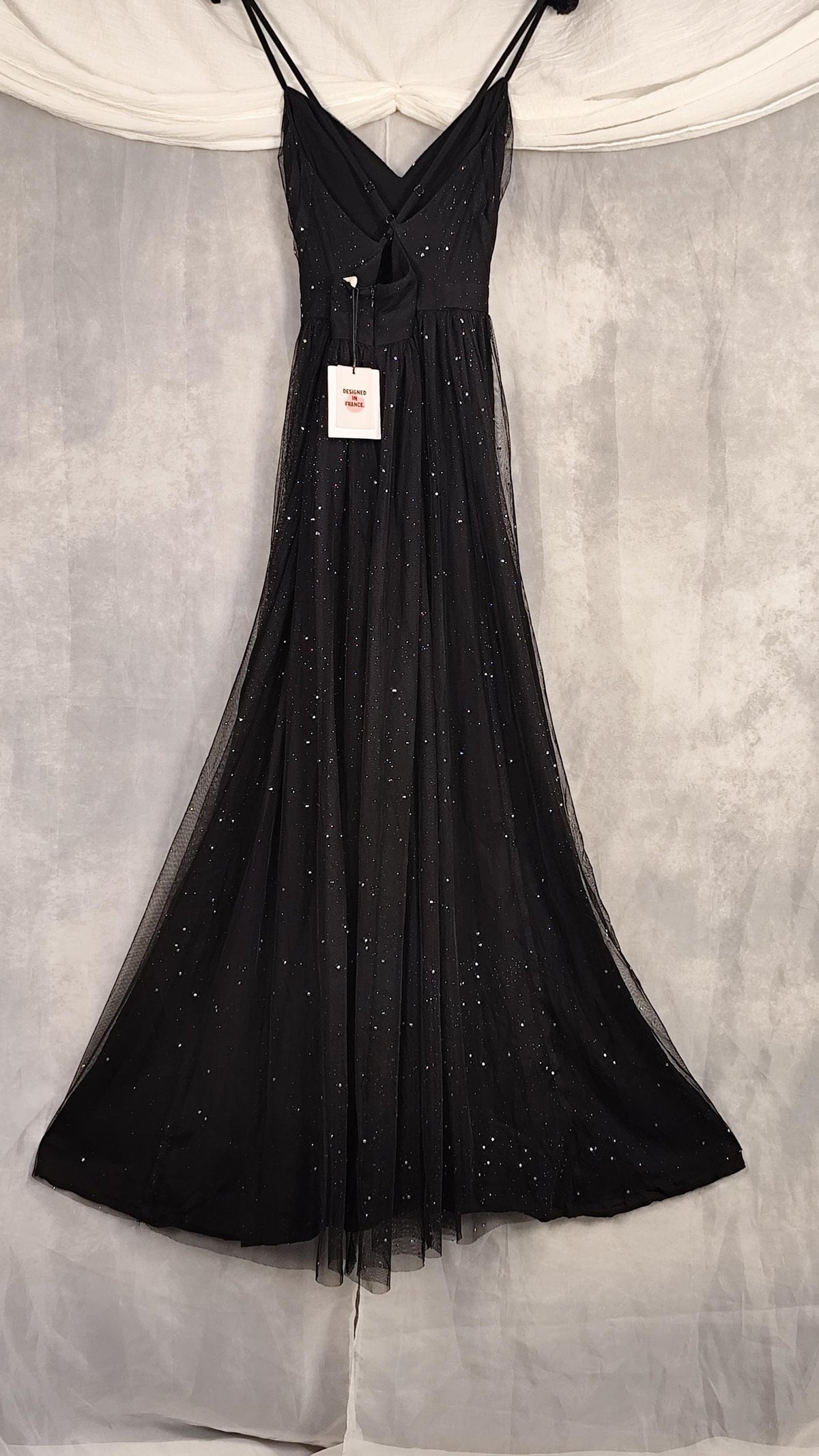 A black sleeveless evening gown with thin spaghetti straps, a V-neckline, and subtle sparkly embellishments, hanging against a gray fabric backdrop with a white draped cloth at the top.