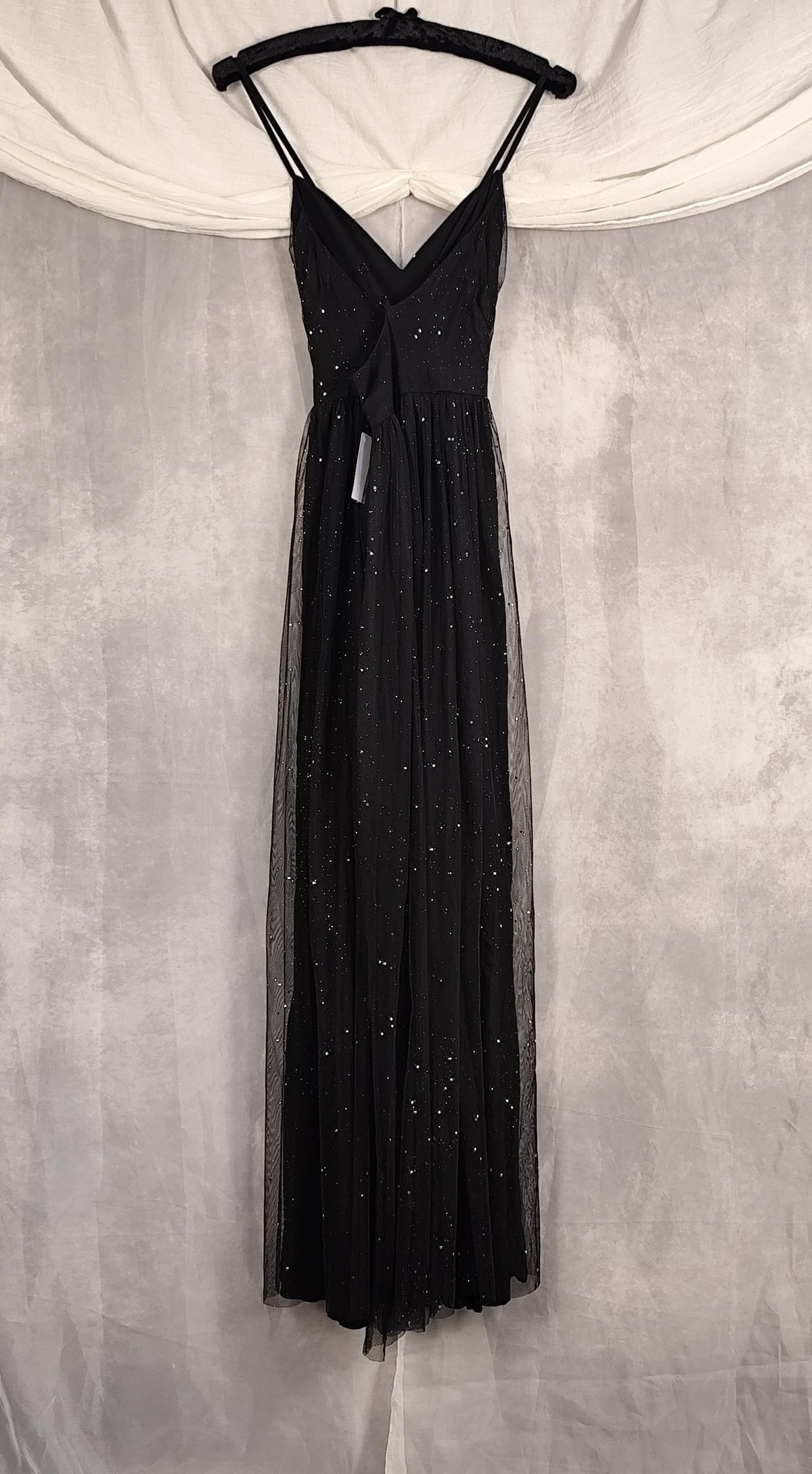 A black sleeveless evening gown with thin straps, a V-neckline, and a sheer tulle skirt, hanging on a black hanger against a gray textured backdrop with white draped fabric at the top.