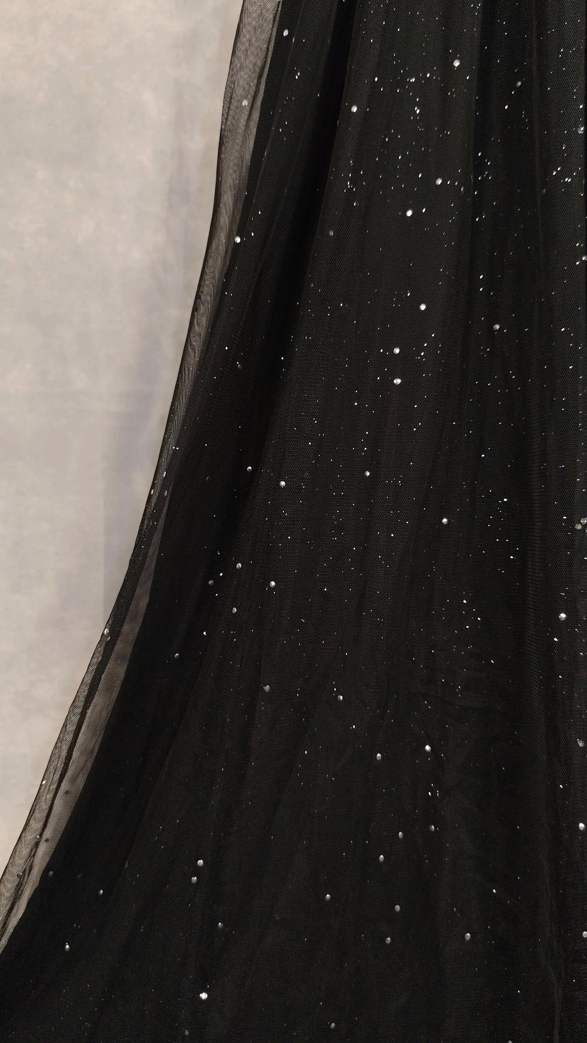 Close-up of a black sheer tulle fabric with scattered small silver glitter or sequin speckles, draped against a light gray background.