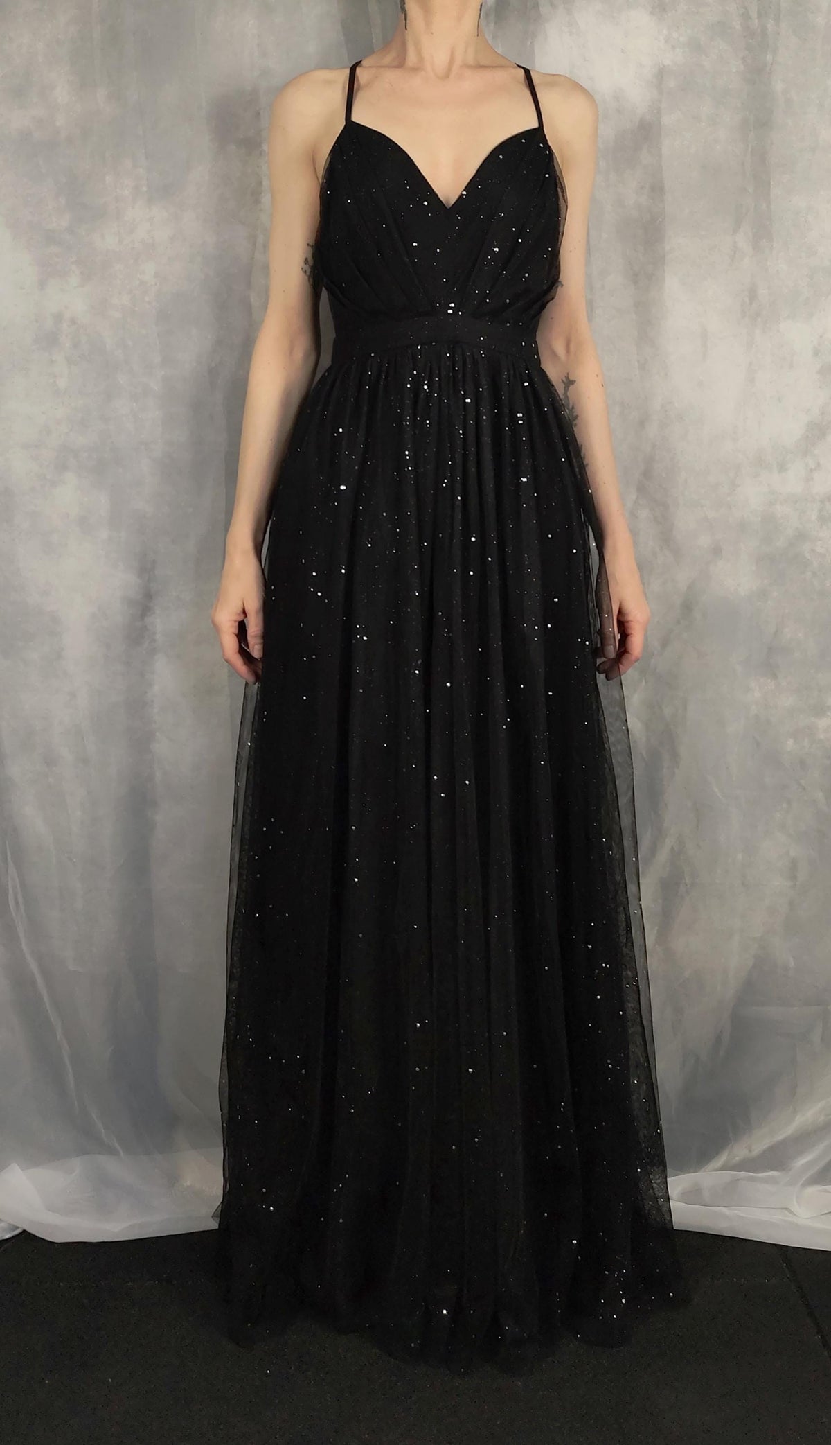 Front view of a black spaghetti-strap evening gown with a sweetheart neckline, fitted bodice, and a full tulle skirt adorned with sequins, shown on a model against a gray backdrop.