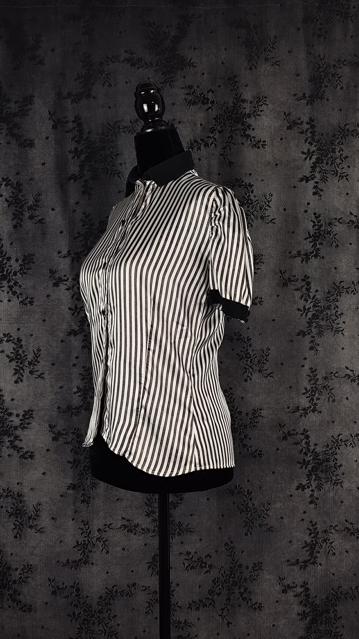 A side view of a black and white vertically striped short-sleeve button-up shirt displayed on a black dress form mannequin against a dark patterned backdrop.