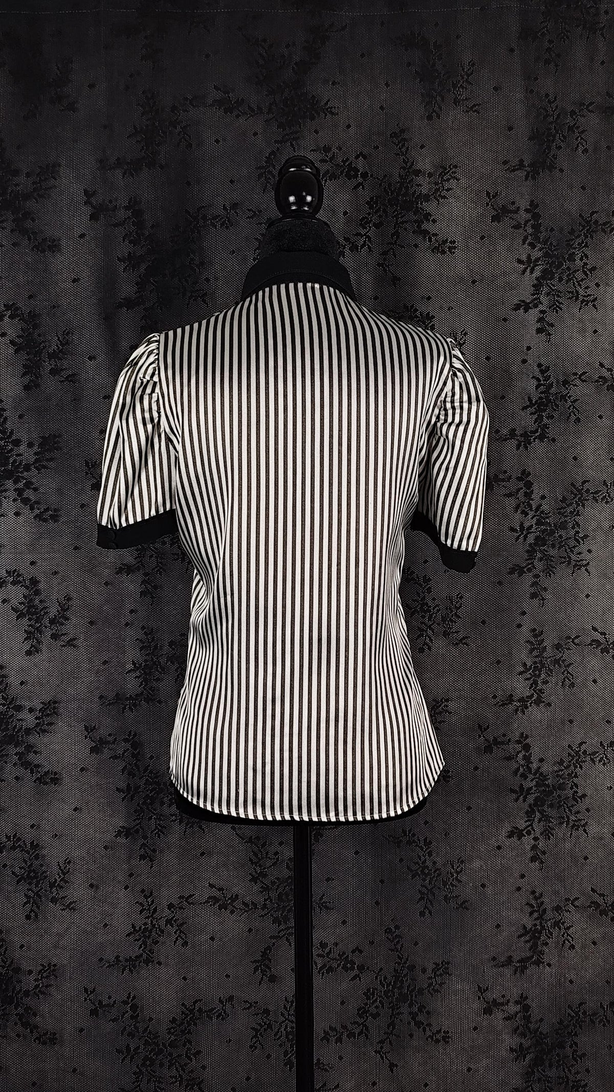 Back view of a black and white vertically striped short-sleeve blazer displayed on a black mannequin torso against a dark textured backdrop.