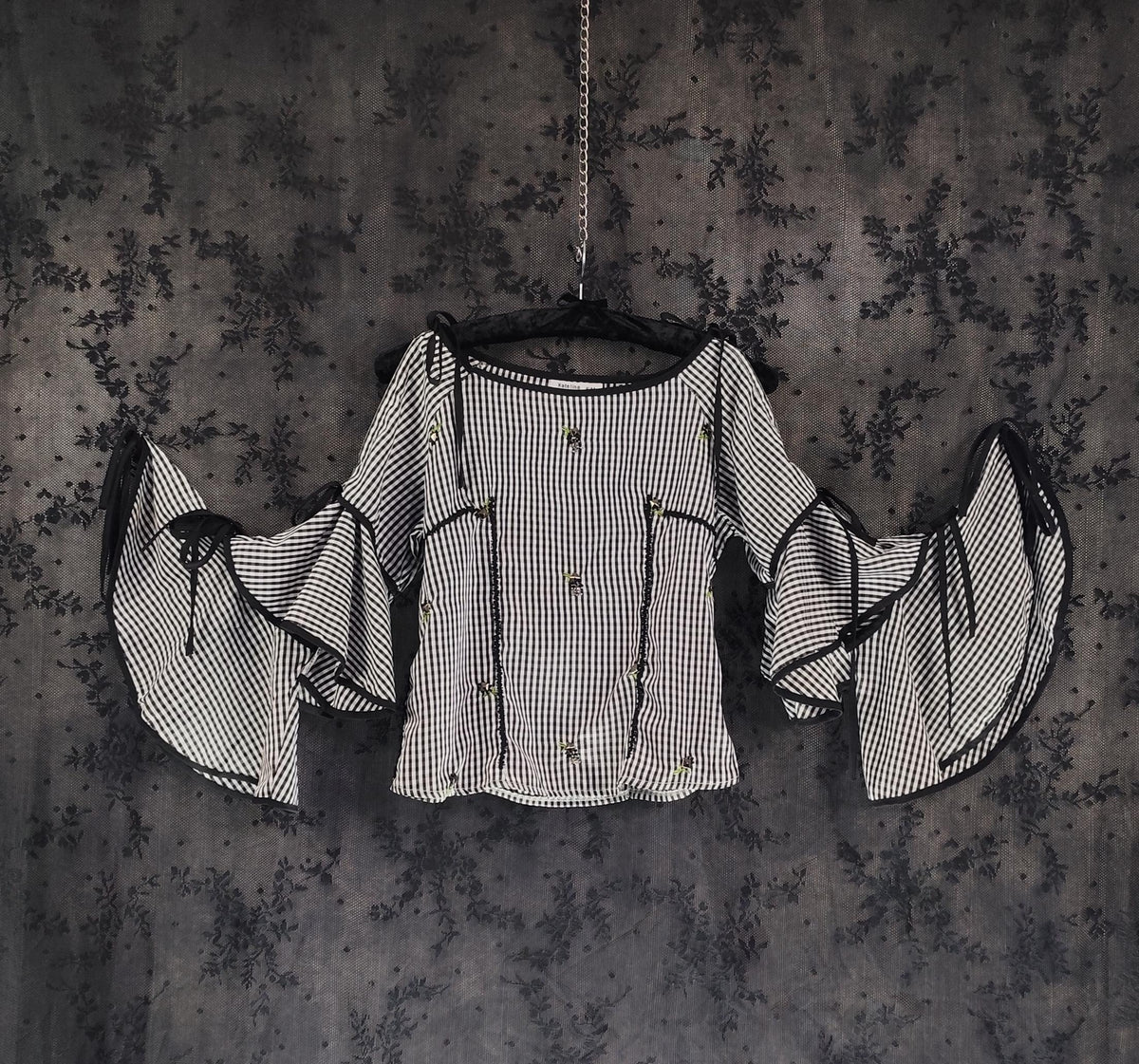 Front view of a black and white checkered blouse with dramatic bell sleeves and black trim, hanging on a black hanger against a dark textured background.