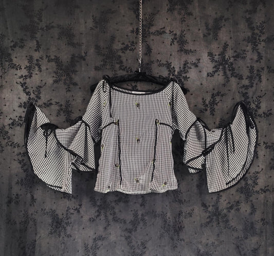 Front view of a black and white checkered blouse with dramatic bell sleeves and black trim, hanging on a black hanger against a dark textured background.