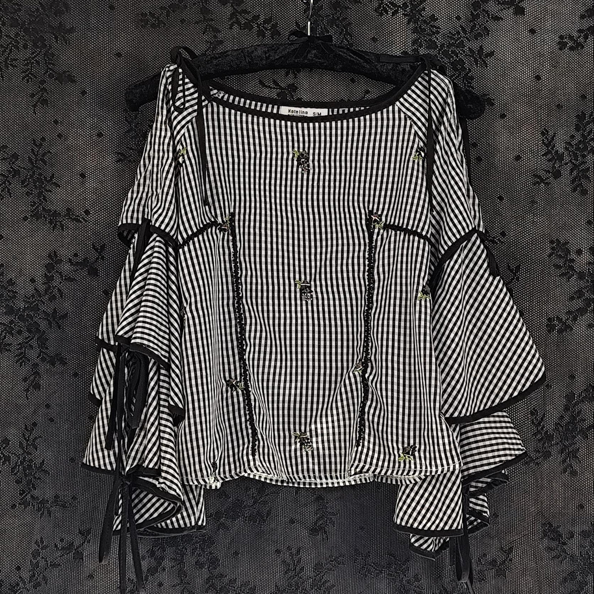 A black and white checkered blouse with layered ruffle sleeves and tie details, hanging on a black hanger against a dark floral wallpaper background.