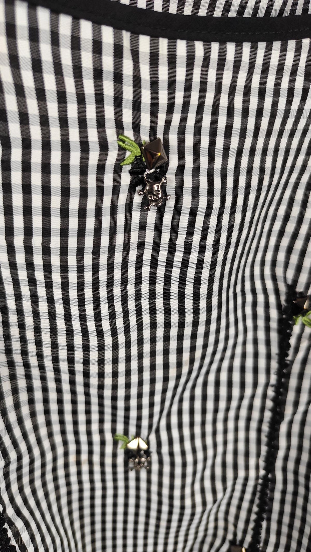 Close-up of a black and white checkered blouse decorated with black ruffled trim, small green floral accents, studs and skulls.