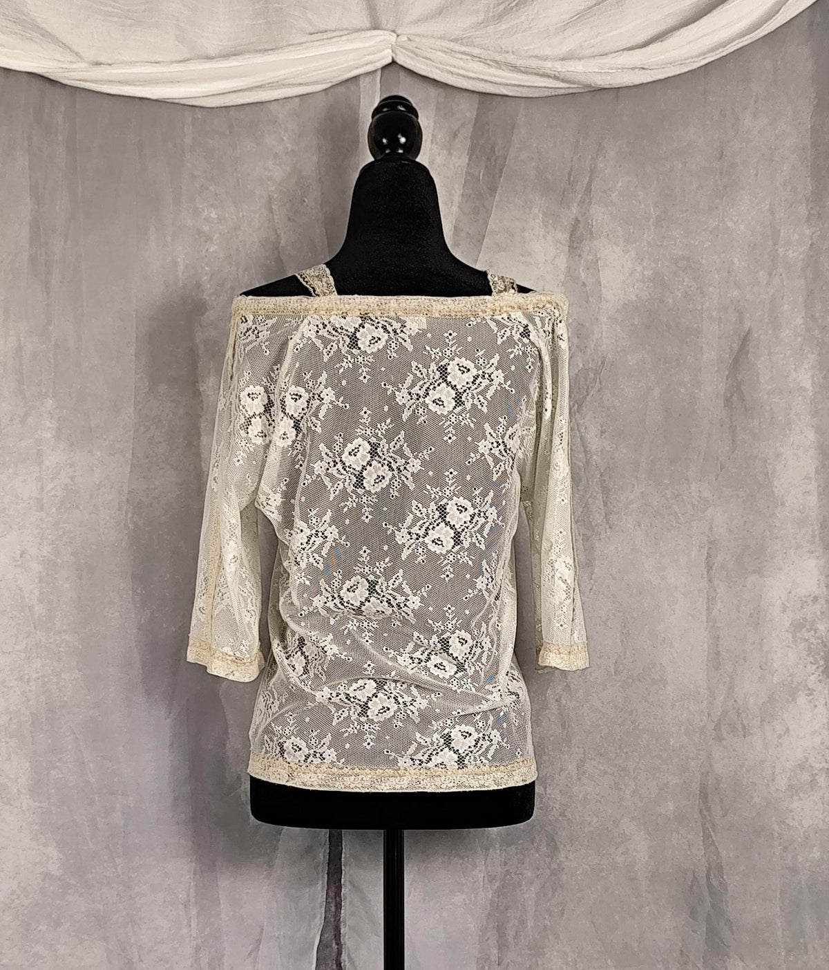 Back view of an off-the-shoulder lace top in cream and pearl, displayed on a black dress form mannequin against a textured gray backdrop with draped white fabric at the top.