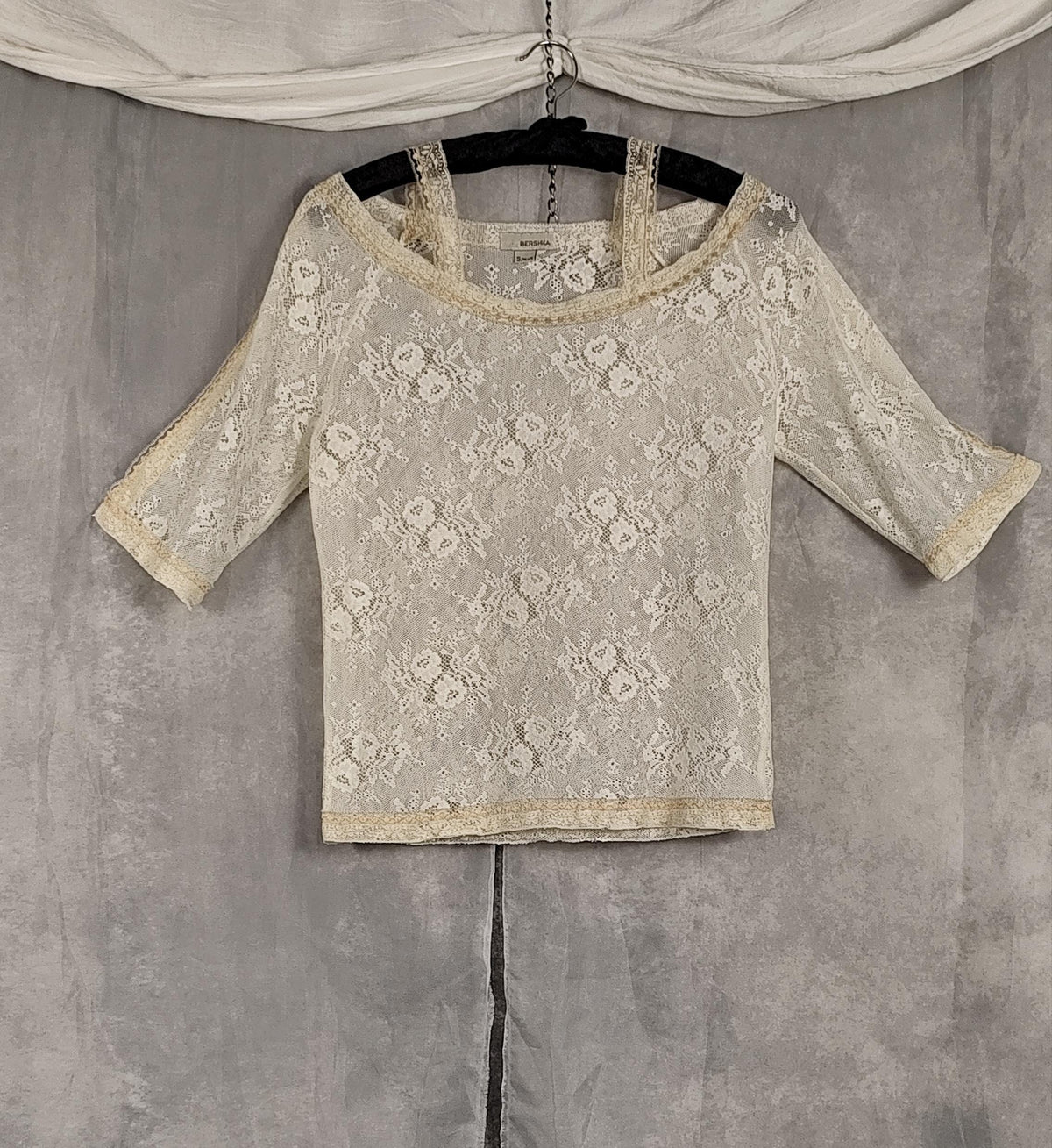 Front view of a pearl-colored lace 3/4 sleeve top with a floral pattern and lace trim, hanging on a black hanger against a textured gray backdrop.