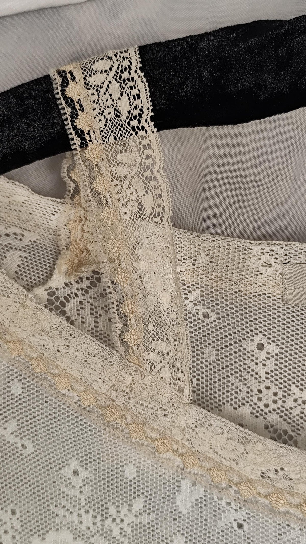 Close-up of a sheer lace top in pearl, showing floral lace patterns and trim, draped over a black hanger against a soft gray background.