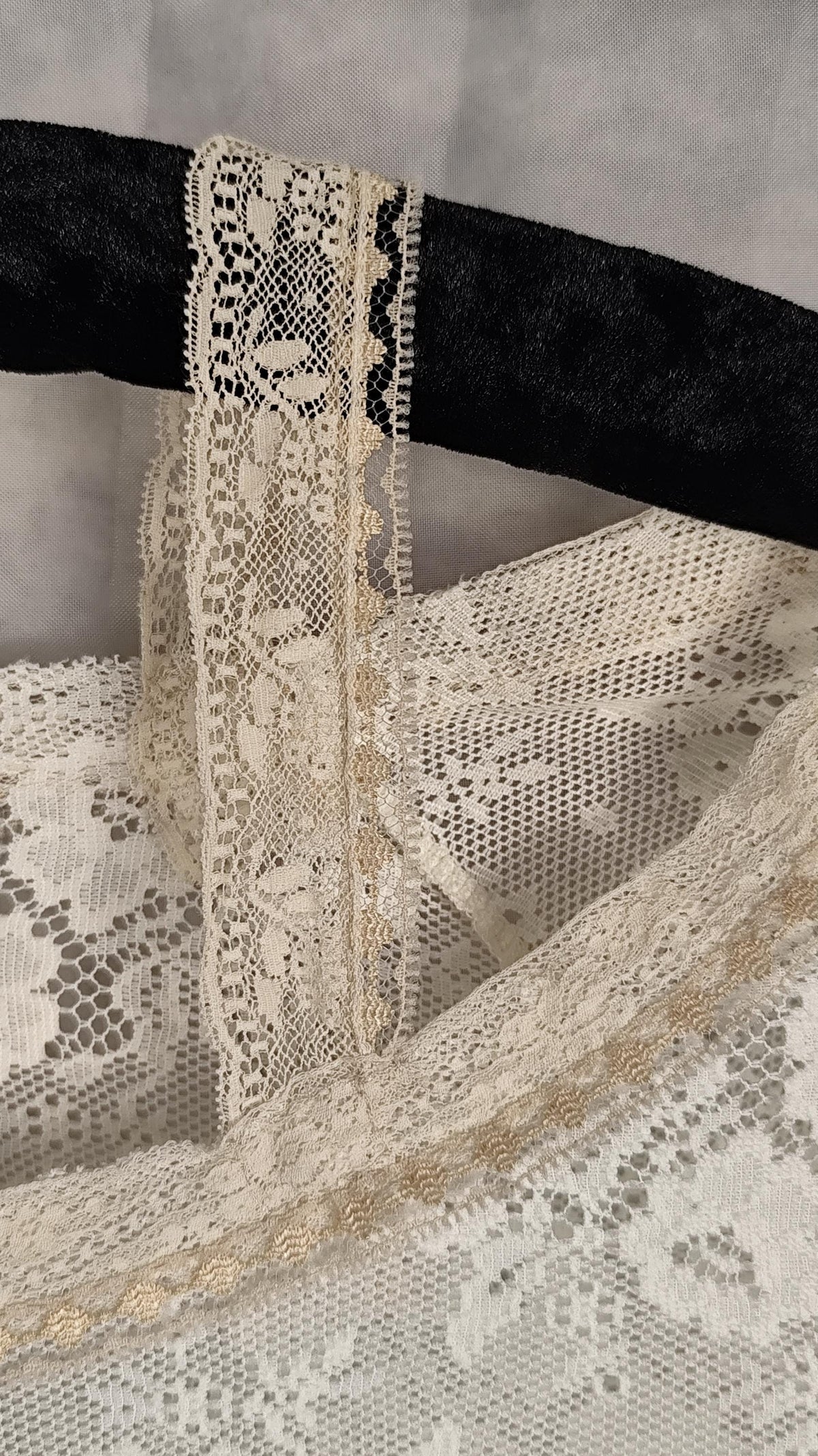 Close-up of a sheer lace top in pearl, showing floral lace patterns and trim, draped over a black hanger against a soft gray background.
