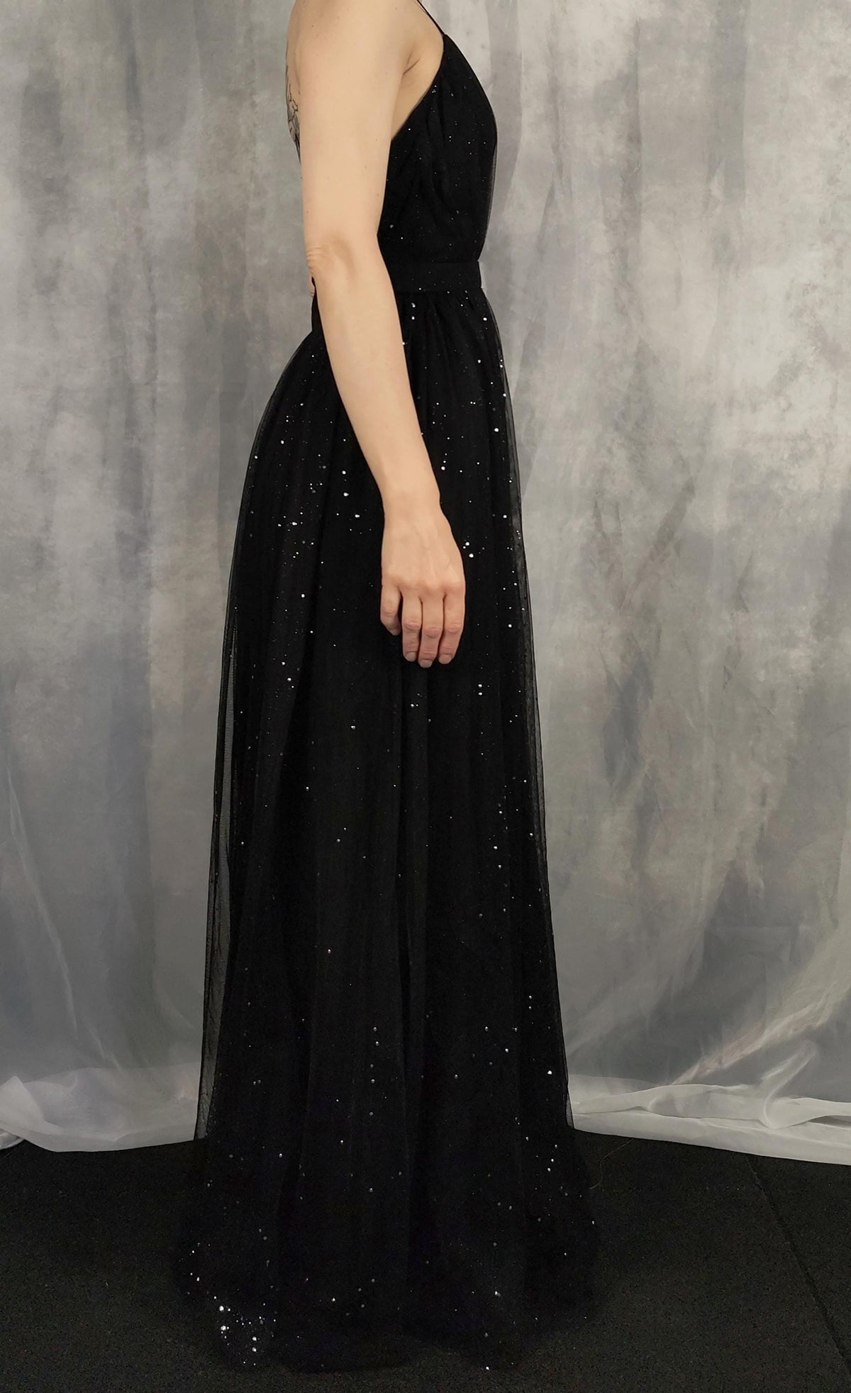 A woman wearing a sleeveless black tulle evening gown with scattered sequins, shown from a side-back angle against a gray textured backdrop.