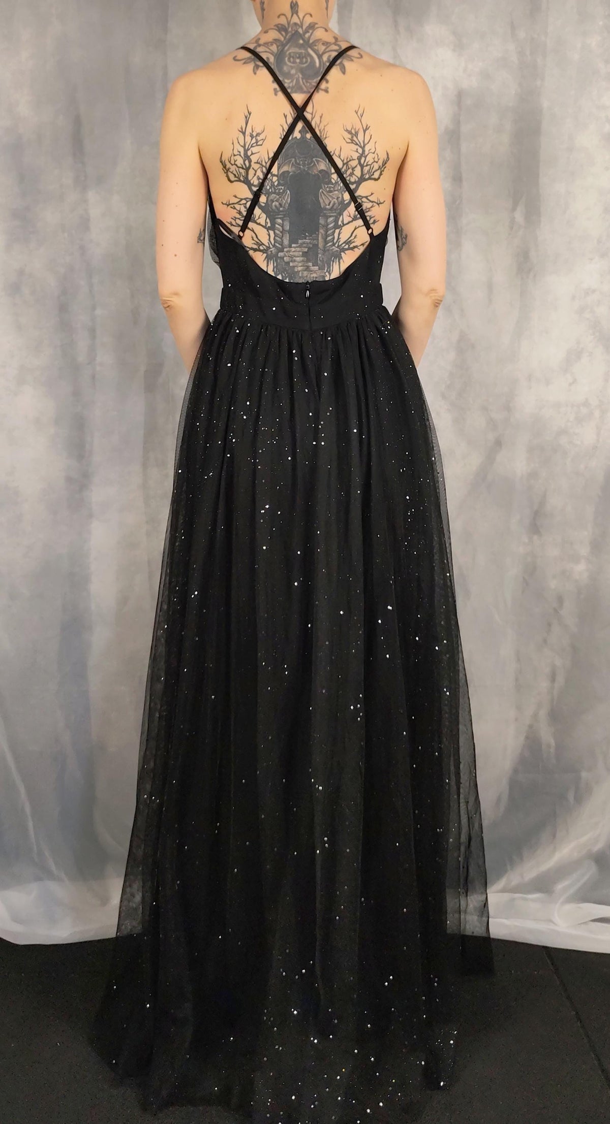 A woman shown from the back wearing a black sleeveless evening gown with an open back, crisscross straps, and a full tulle skirt with scattered rhinestone embellishments, posed against a gray studio backdrop.